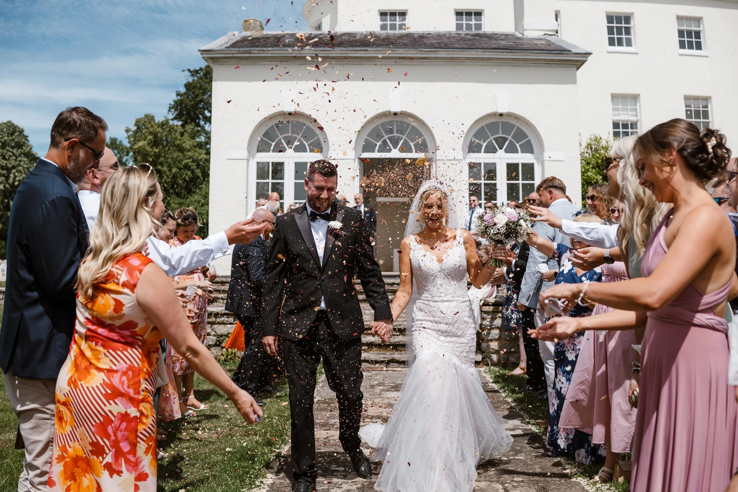 A newlywed couple holding hands walking away from an outdoor wedding ceremony as guests throw confetti in the air on a sunny day.