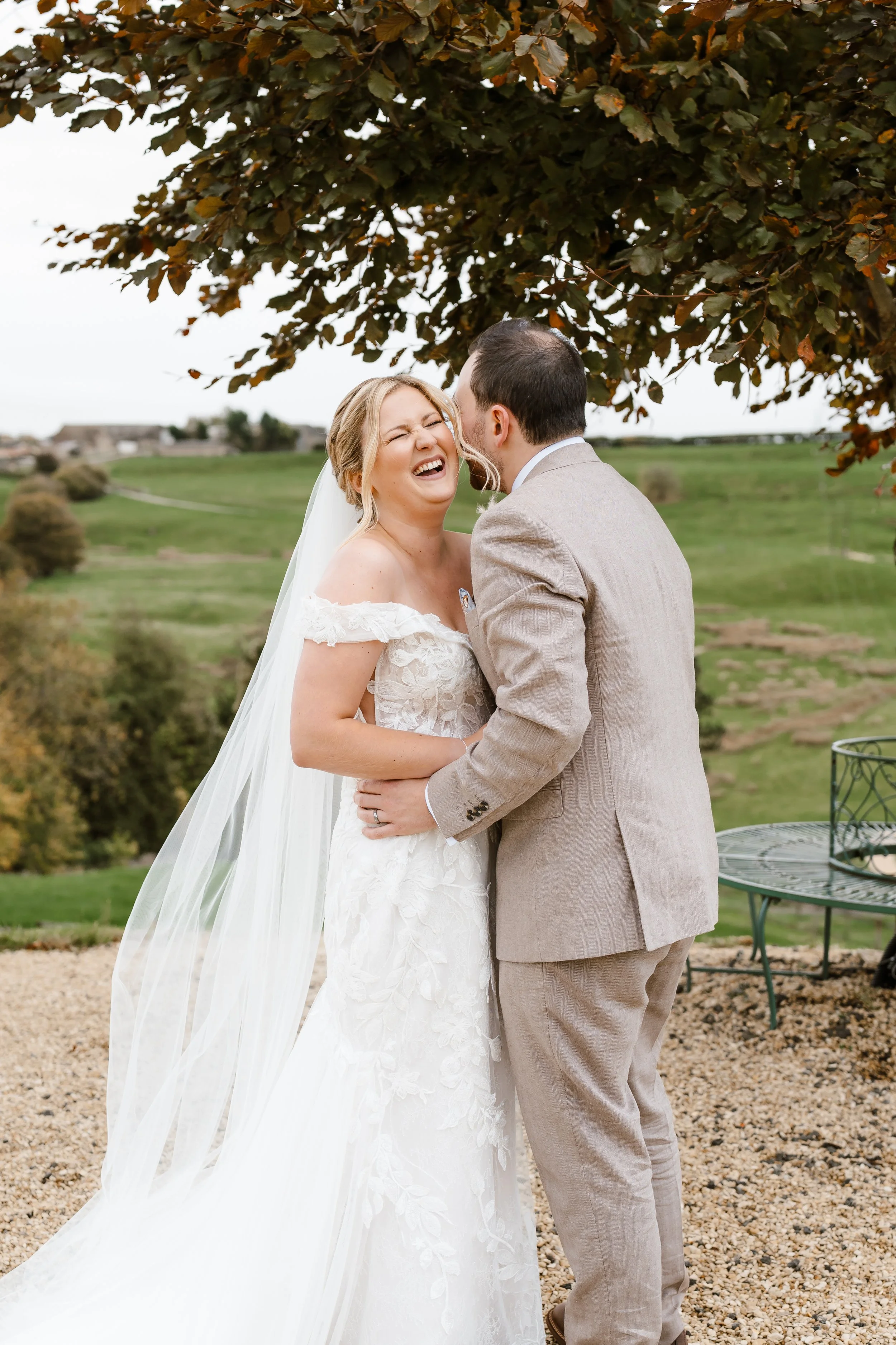 A bride and groom sharing a joyful moment outdoors on their wedding day, with the bride laughing and the groom smiling, standing under a large tree with a scenic landscape in the background.