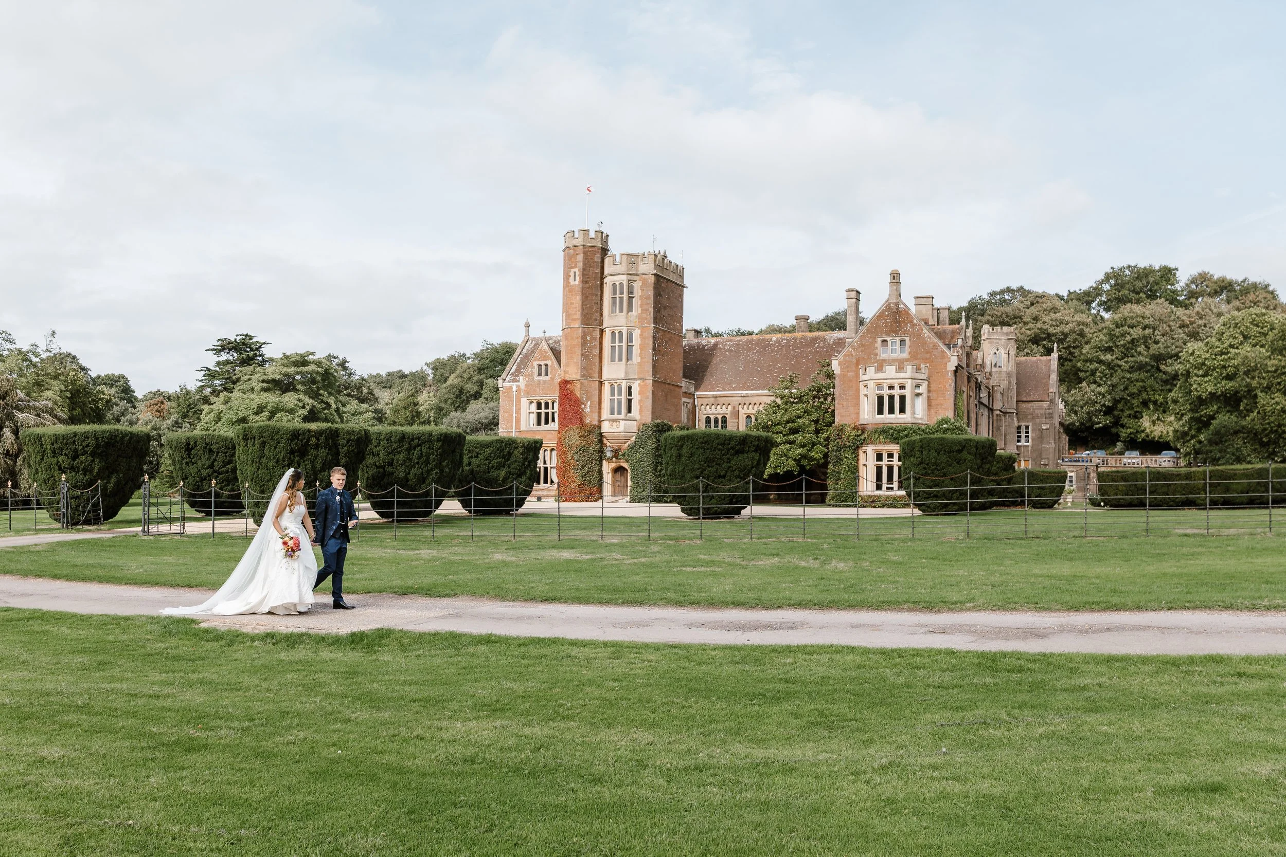 A bride and groom walking on a gravel path in front of a large historic castle with a lush green lawn and trees in the background.