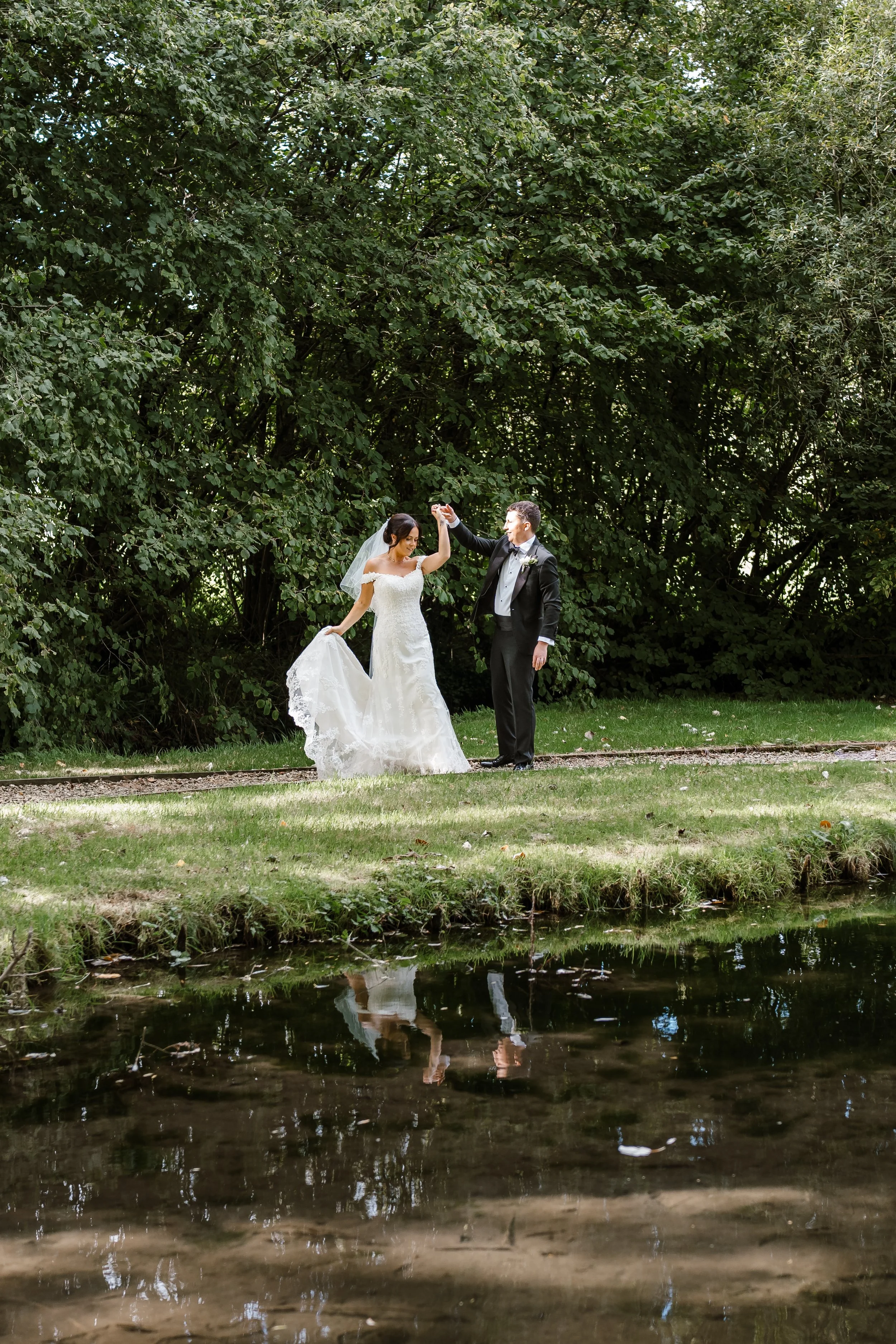 A bride and groom dancing outdoors near a pond with green trees in the background.