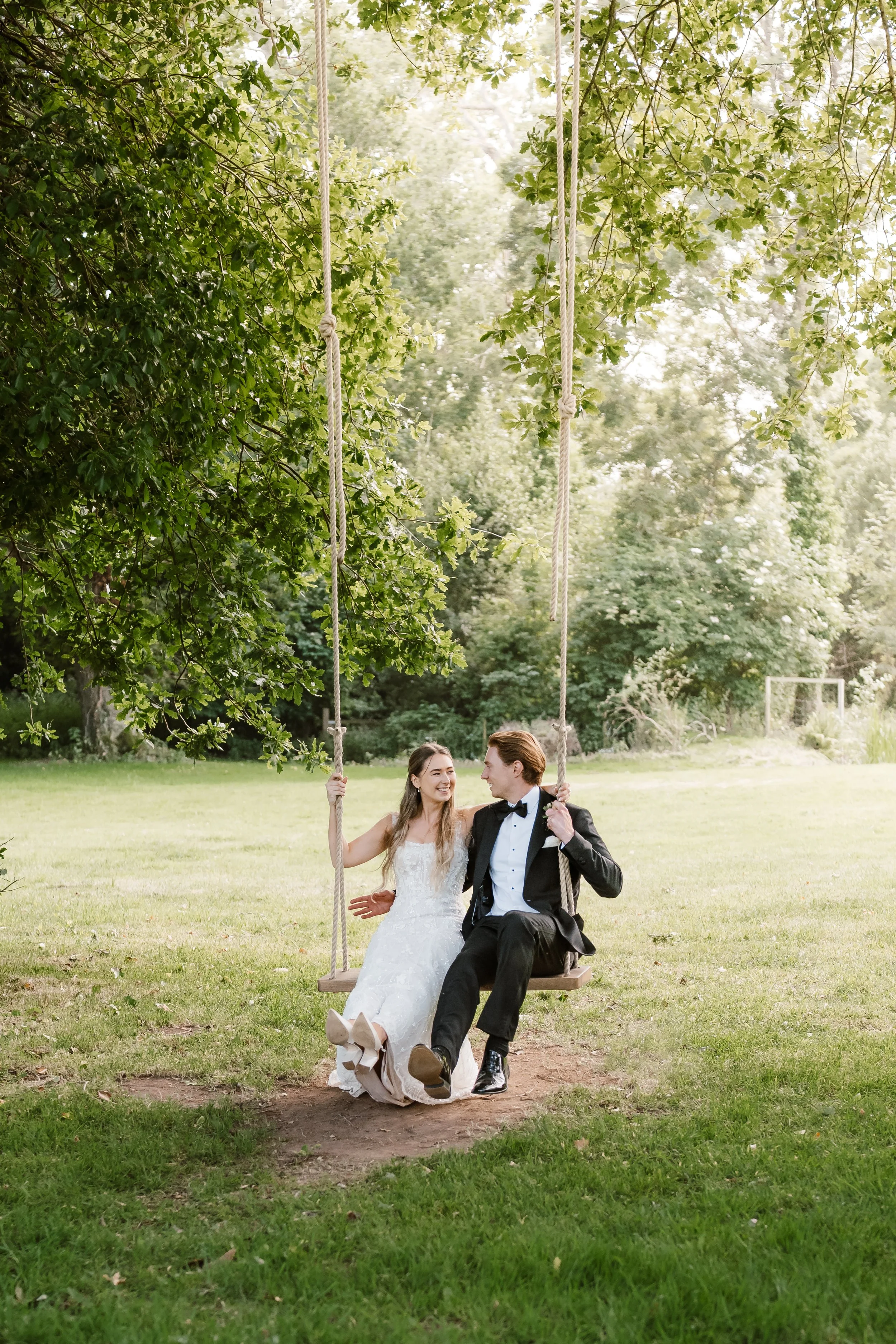 Couple on a wooden swing hanging from a tree, smiling and looking at each other, outdoors in a grassy park with trees in the background, possibly at a wedding or special event.