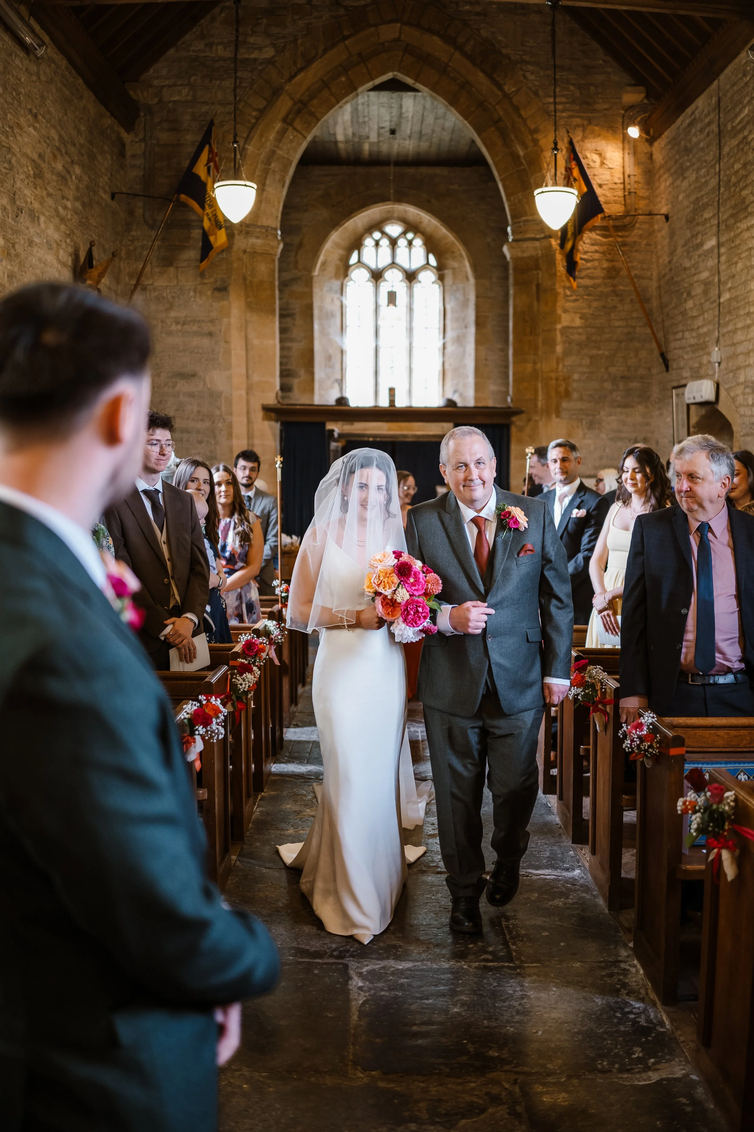 A bride walking down the aisle with her father in a church, surrounded by guests dressed in formal attire, with wooden pews and a large stained glass window at the altar.