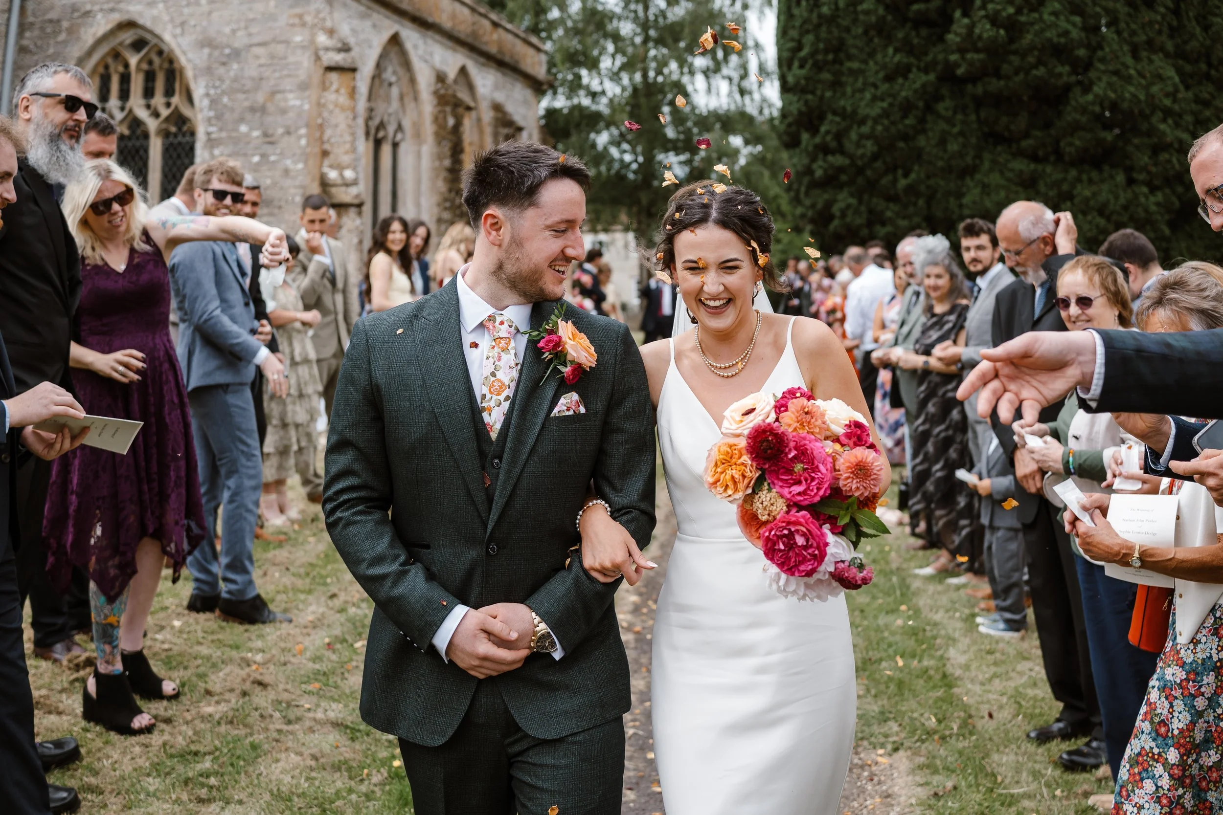 A newlywed couple is walking arm-in-arm outdoors after their wedding ceremony, smiling and laughing. The bride is holding a colorful bouquet of flowers, and the groom is dressed in a dark suit with a floral tie. Guests are gathered around, some throw