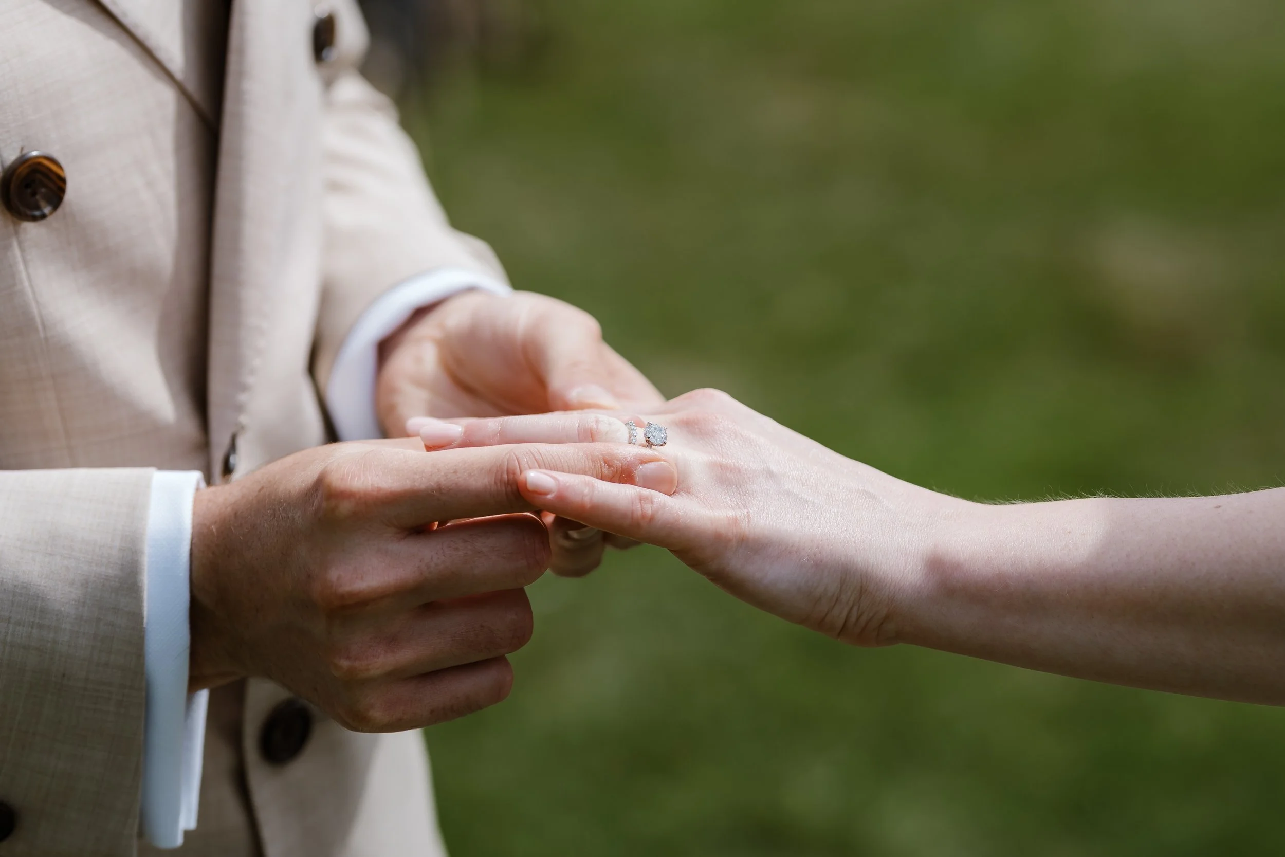 A person wearing a beige blazer with dark buttons is placing a ring on the finger of another person, with a green blurred background.