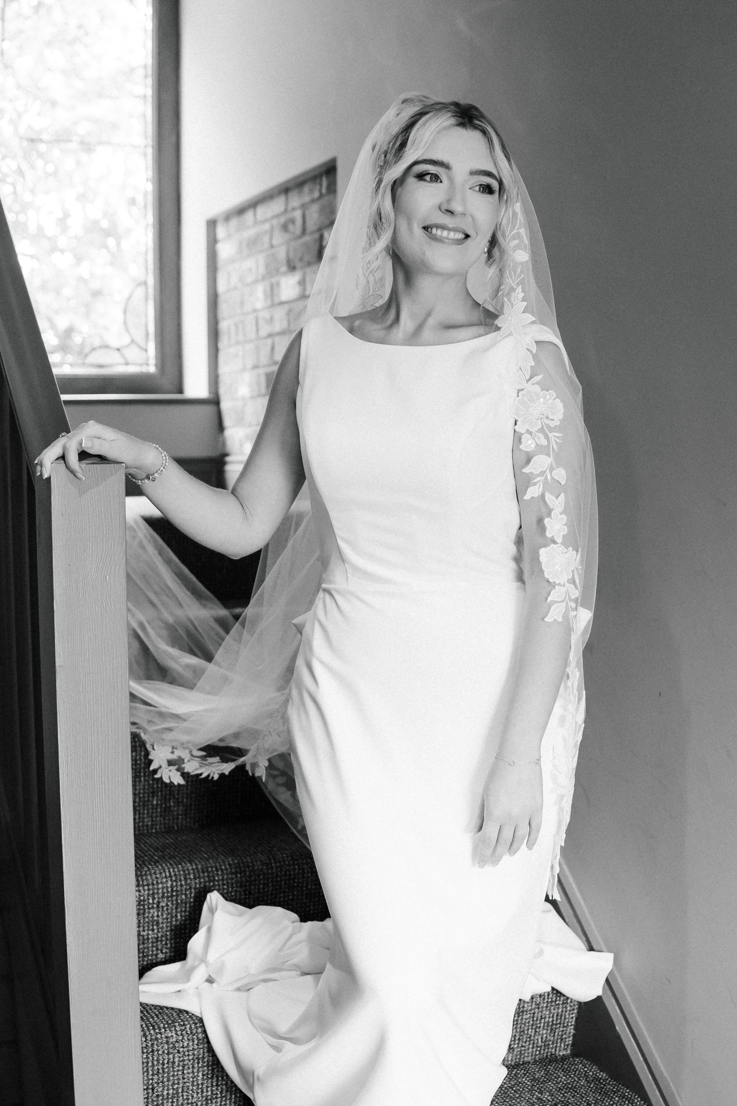 A woman in a wedding dress with a veil, standing on stairs, smiling and looking away. The image is in black and white.