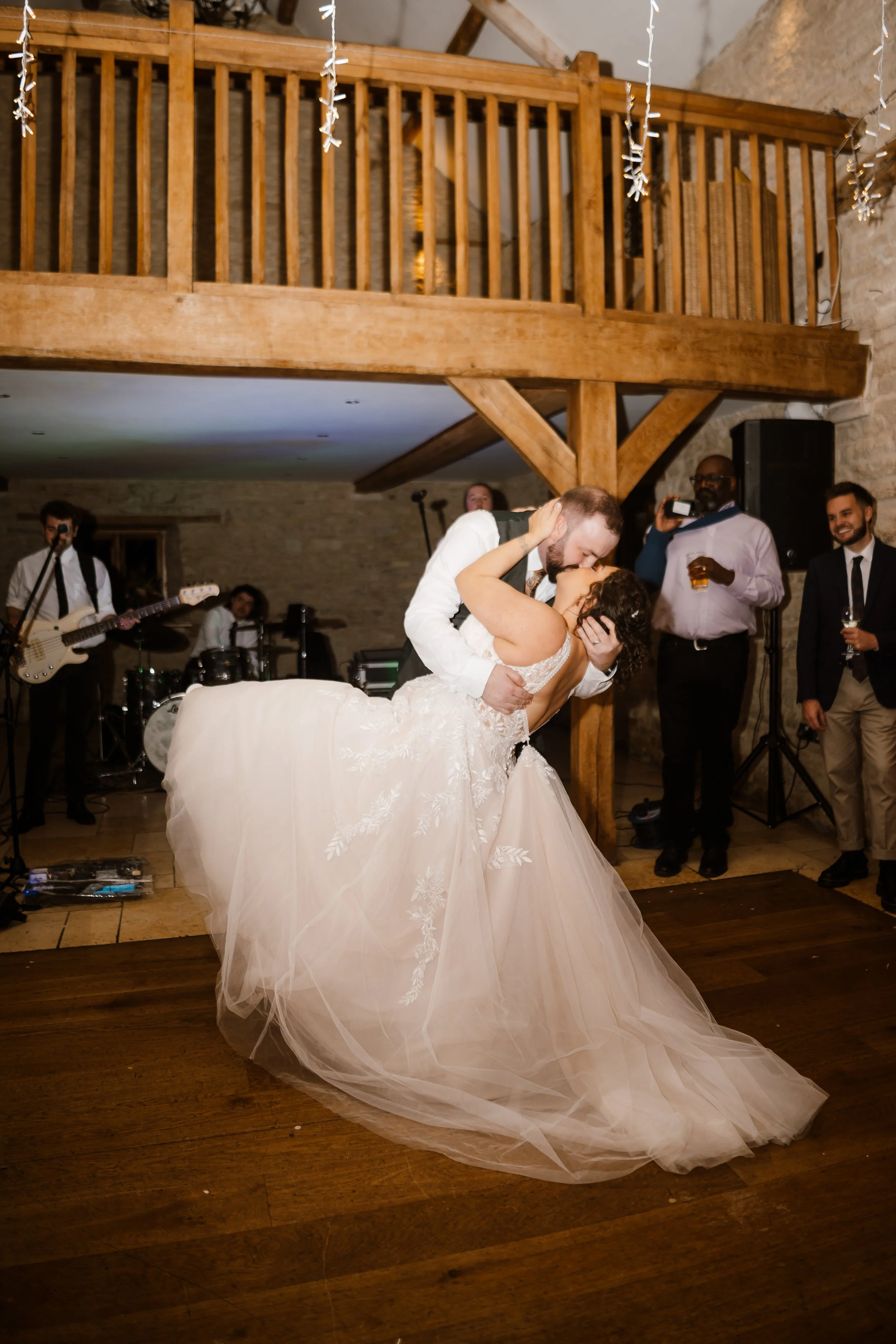 A newlywed couple shares a first dance at their wedding reception, with guests and a live band in the background, in a rustic venue with wooden beams and string lights.
