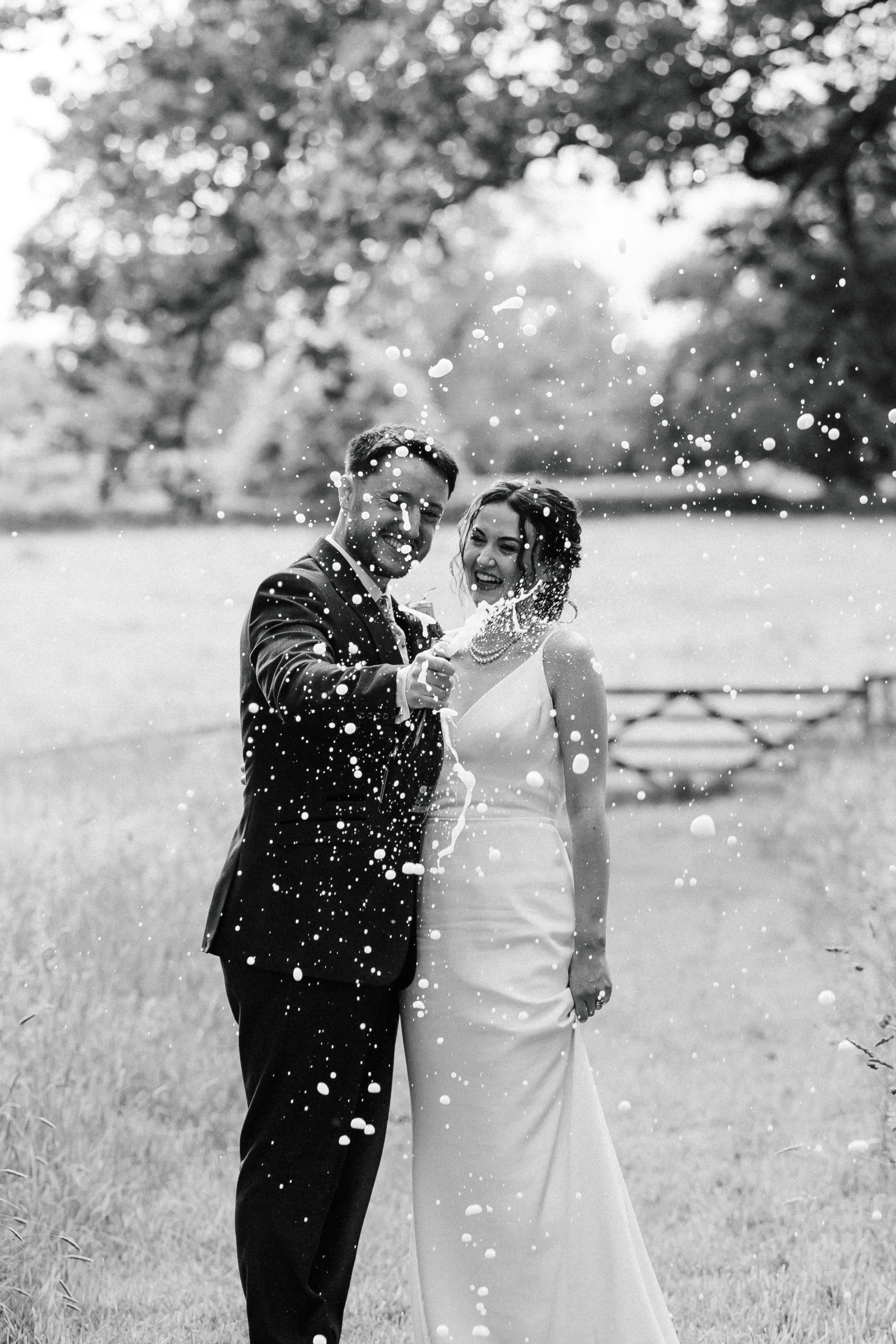 A black-and-white photograph of a newlywed couple celebrating outdoors. The groom is in a suit, and the bride is in a wedding dress. They are playfully spraying water or champagne, with joyful expressions. The background features trees and a grassy field.
