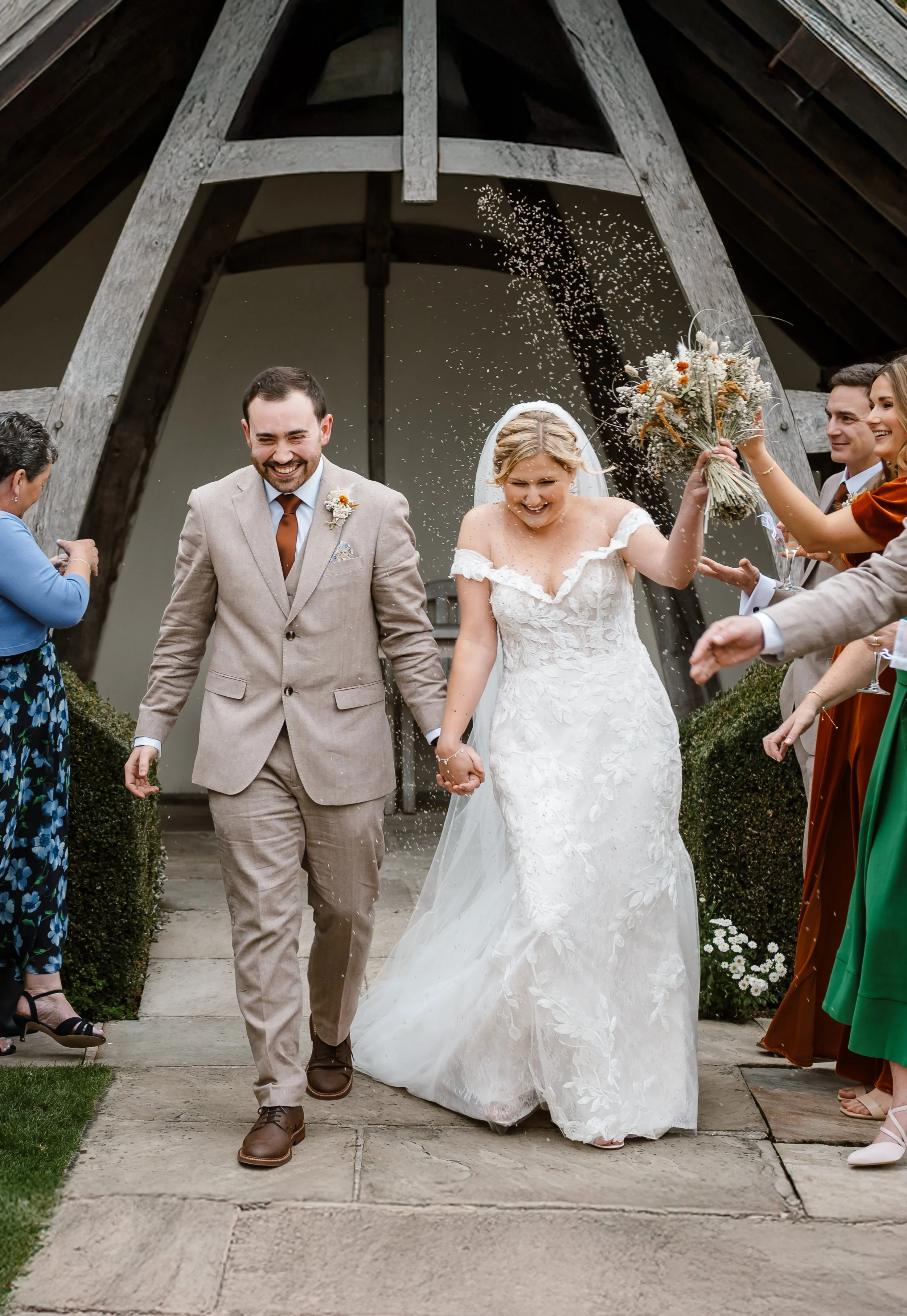 A joyful newlywed couple holding hands and smiling, walking out of a church while guests throw flower petals and celebrate.