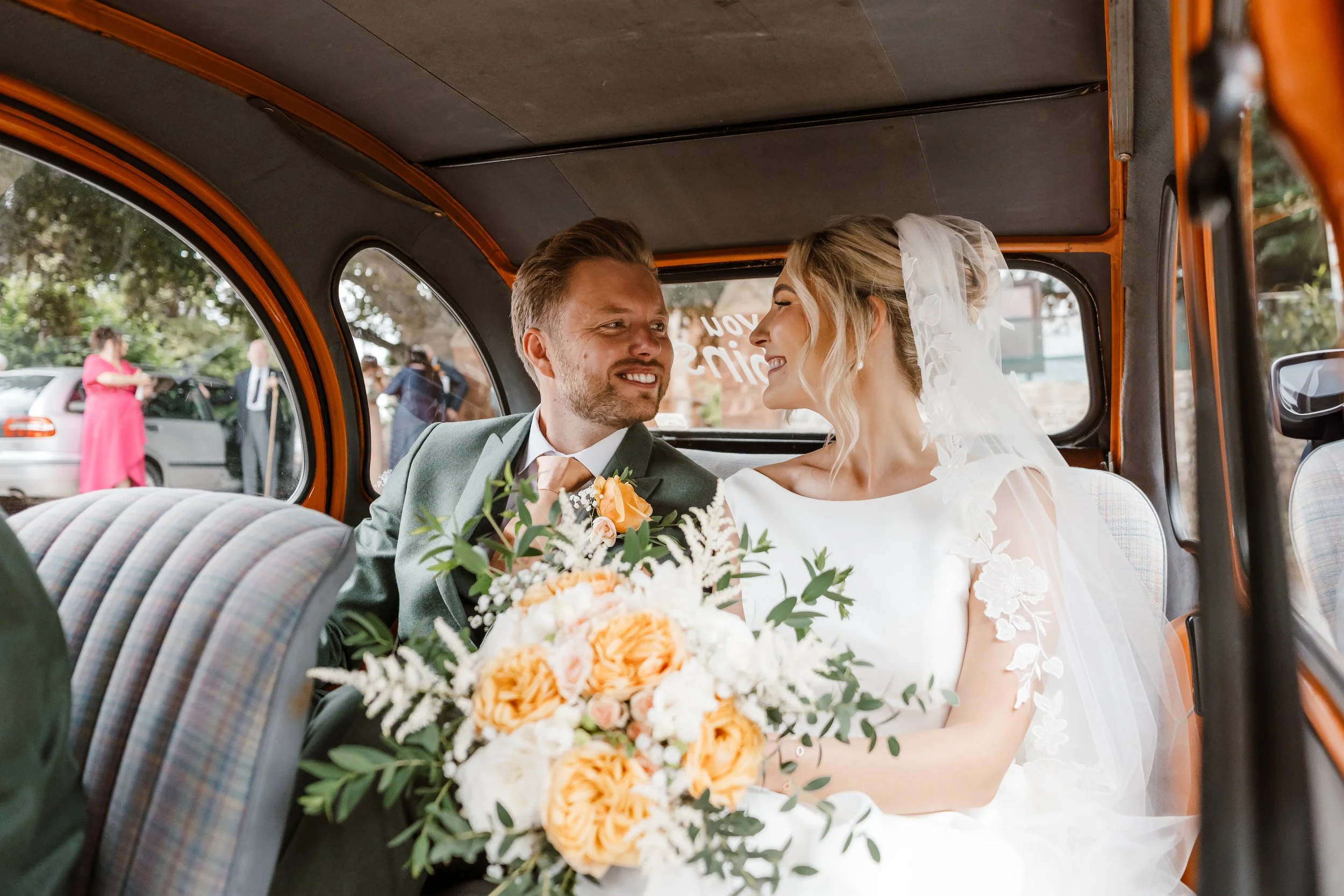 Bride and groom sitting together in a vintage car, smiling at each other; the bride has a bouquet of orange and white flowers.