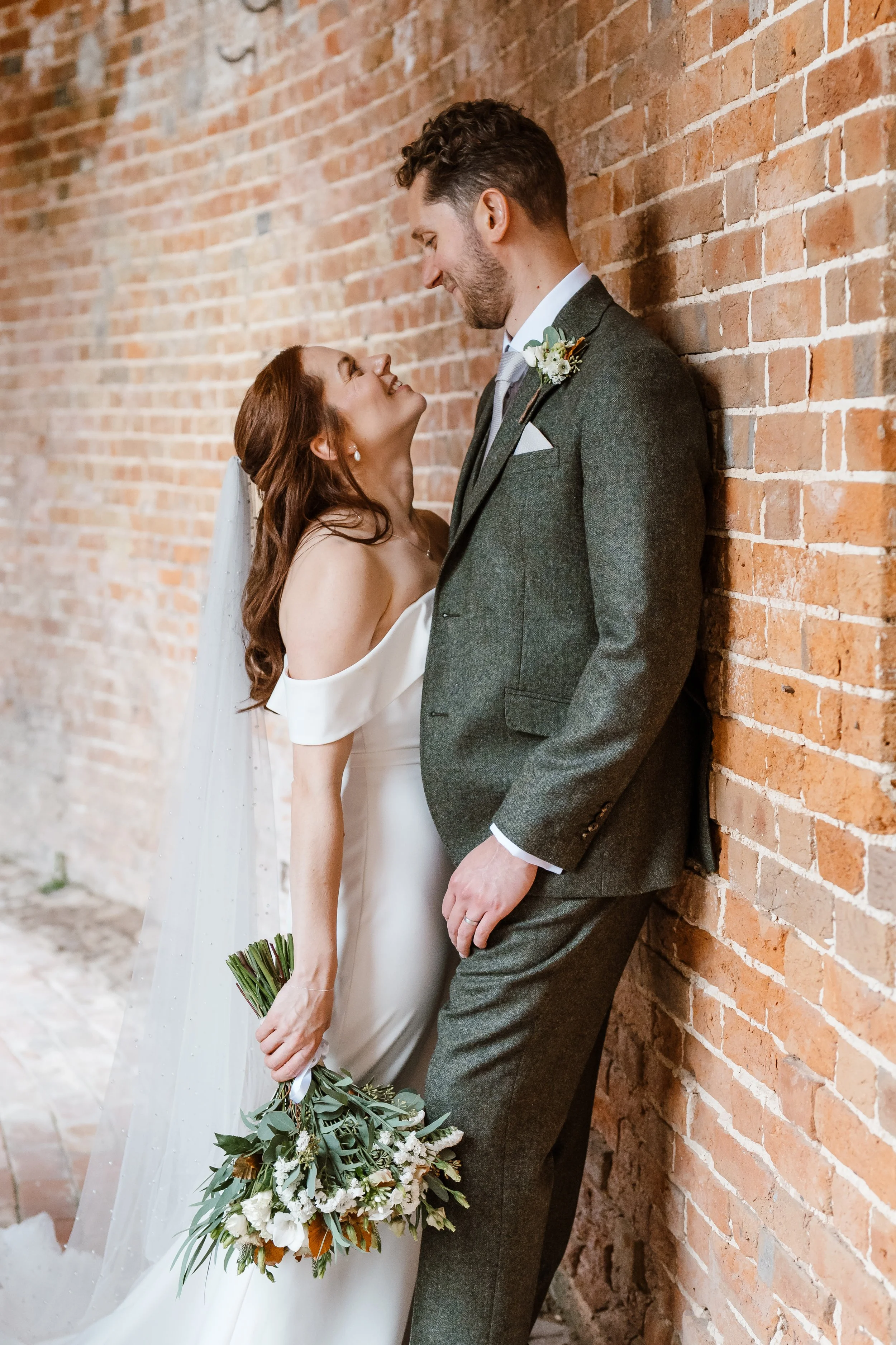 A newlywed couple in wedding attire standing against a brick wall, looking at each other and smiling, with the bride holding a bouquet.