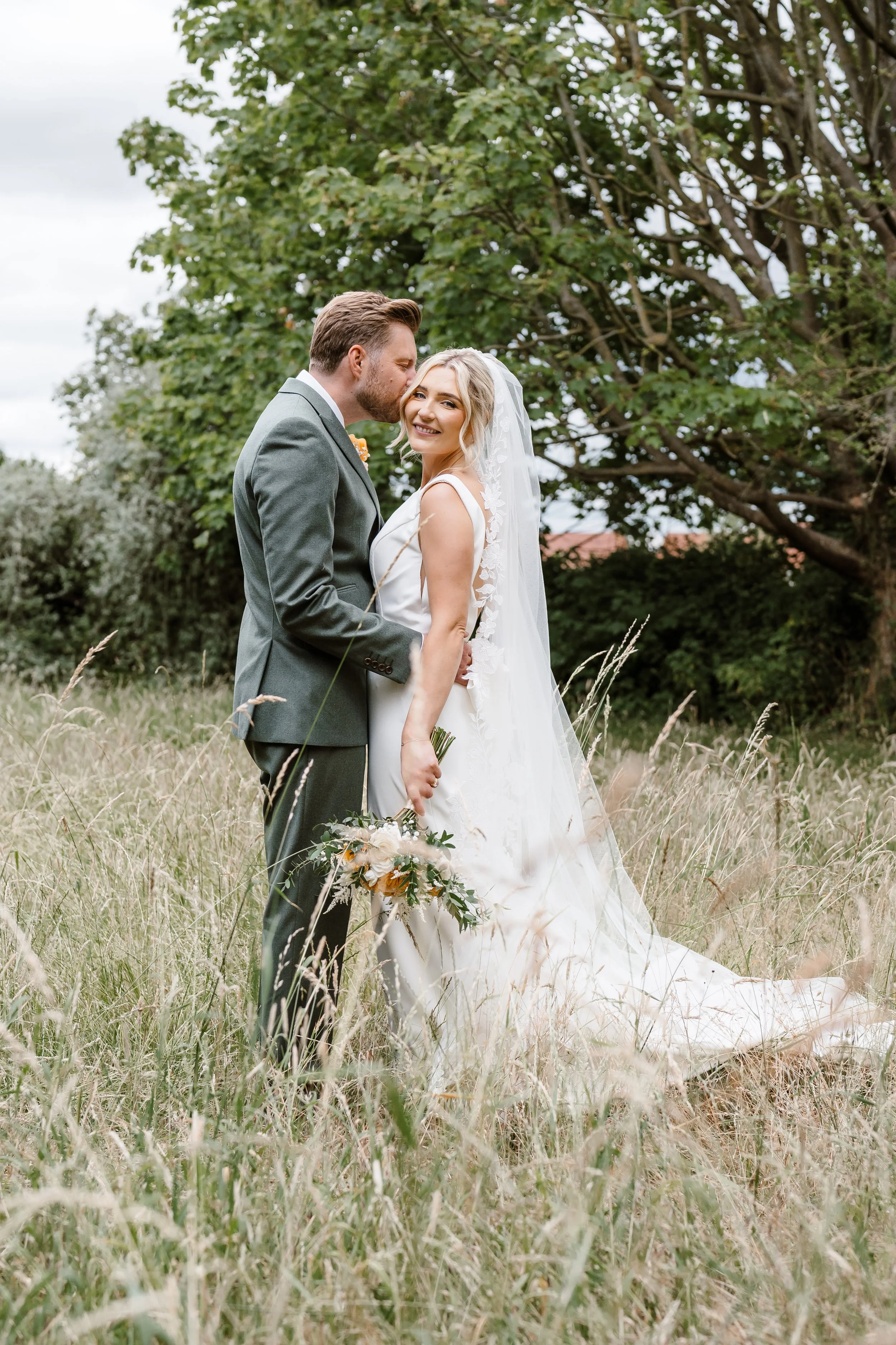 A bride and groom standing closely in a grassy field, with the groom kissing the bride's temple. The bride is holding a bouquet and wearing a white wedding gown with a veil, while the groom is wearing a gray suit. There are trees in the background.
