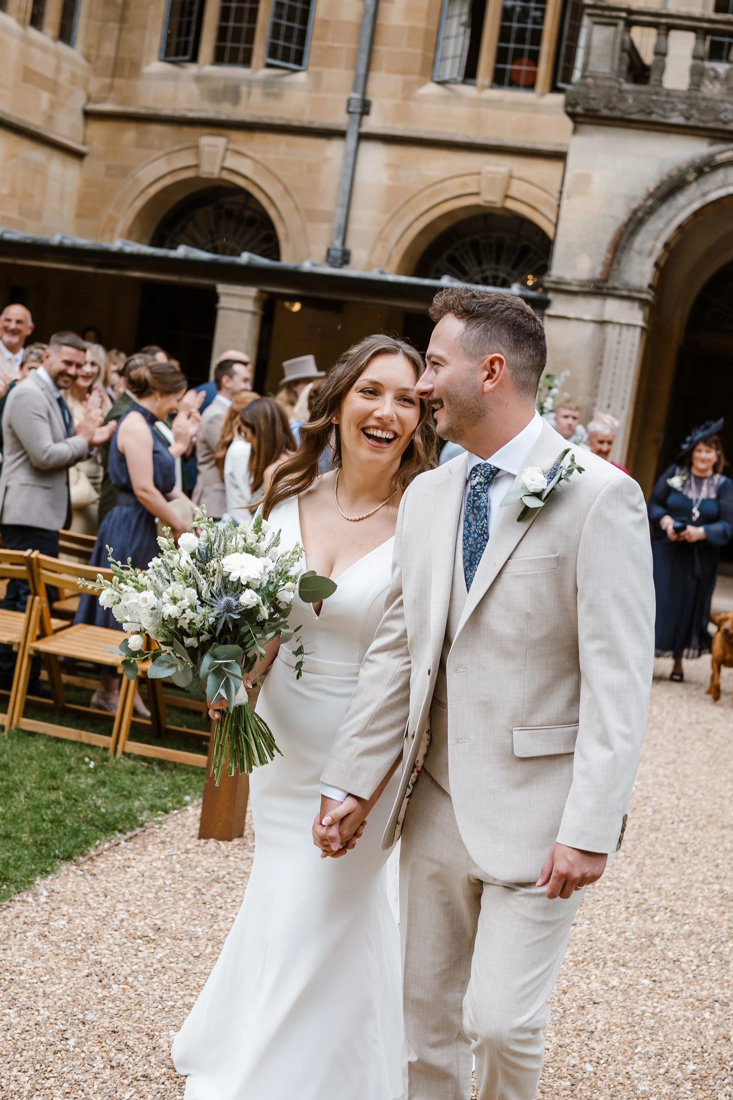A bride and groom walking hand in hand at their wedding ceremony, smiling and looking at each other, in an outdoor courtyard with guests in the background clapping and celebrating.