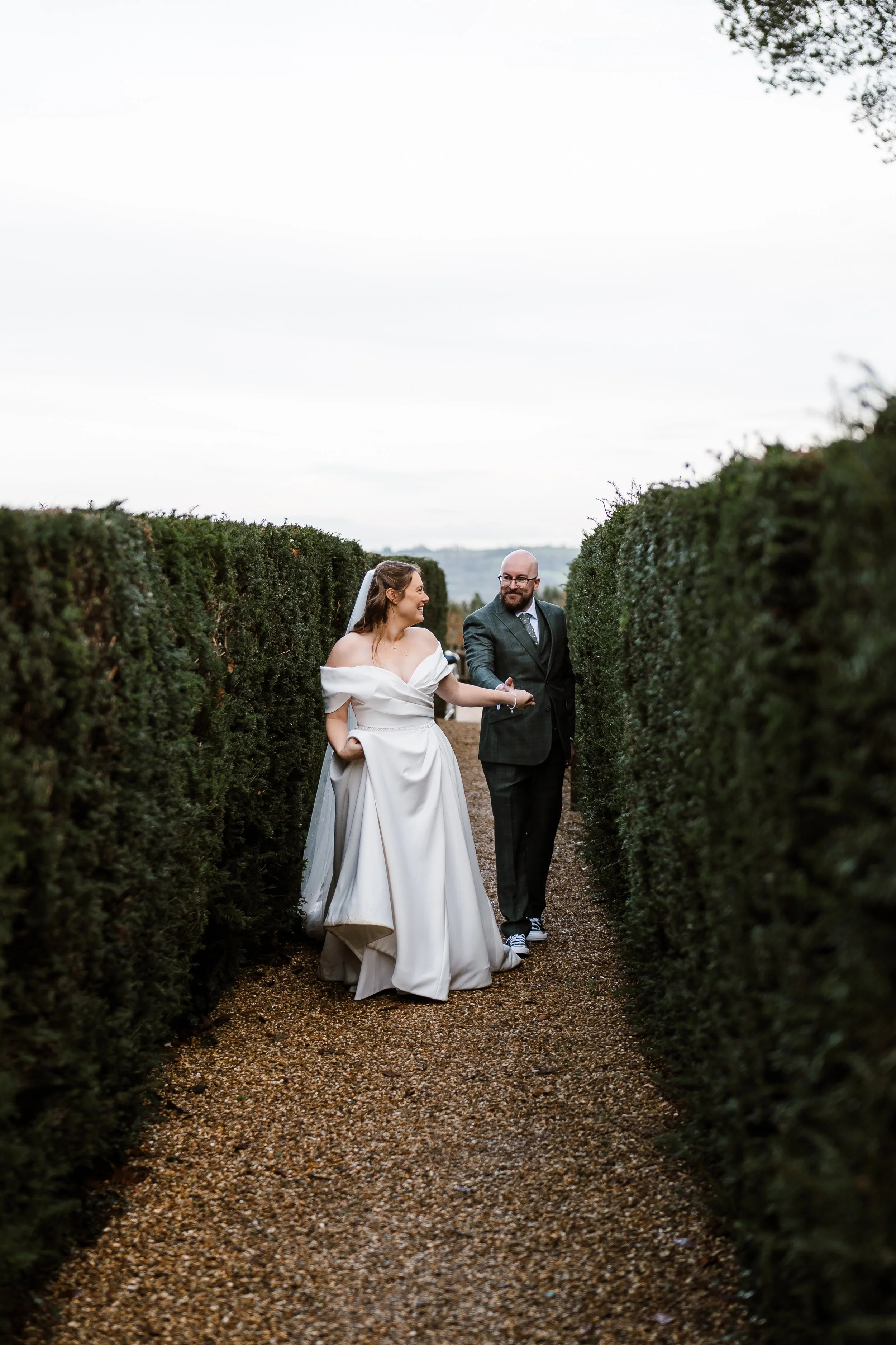 A bride and groom walking hand in hand through a hedge maze, dressed in wedding attire, smiling and enjoying each other's company.