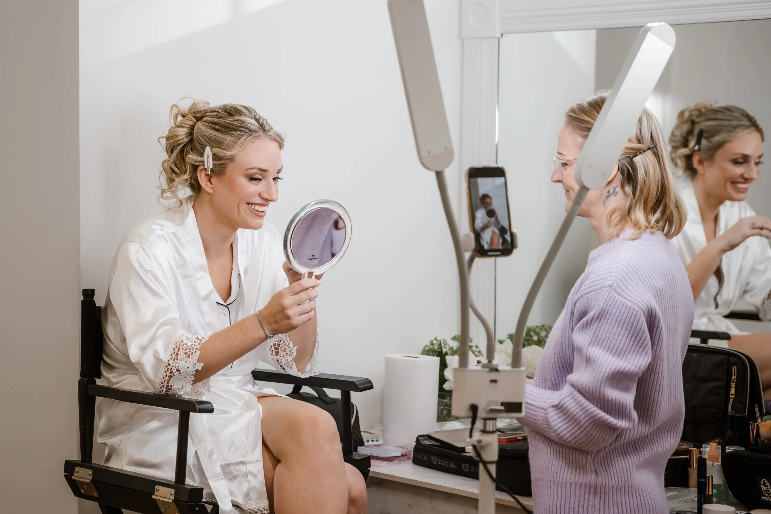 A woman in a white robe sitting in a makeup chair, smiling, while looking into a handheld mirror. She is being filmed or photographed by a woman standing in front of her, who is holding a phone on a selfie stick. The scene appears to be in a makeup o