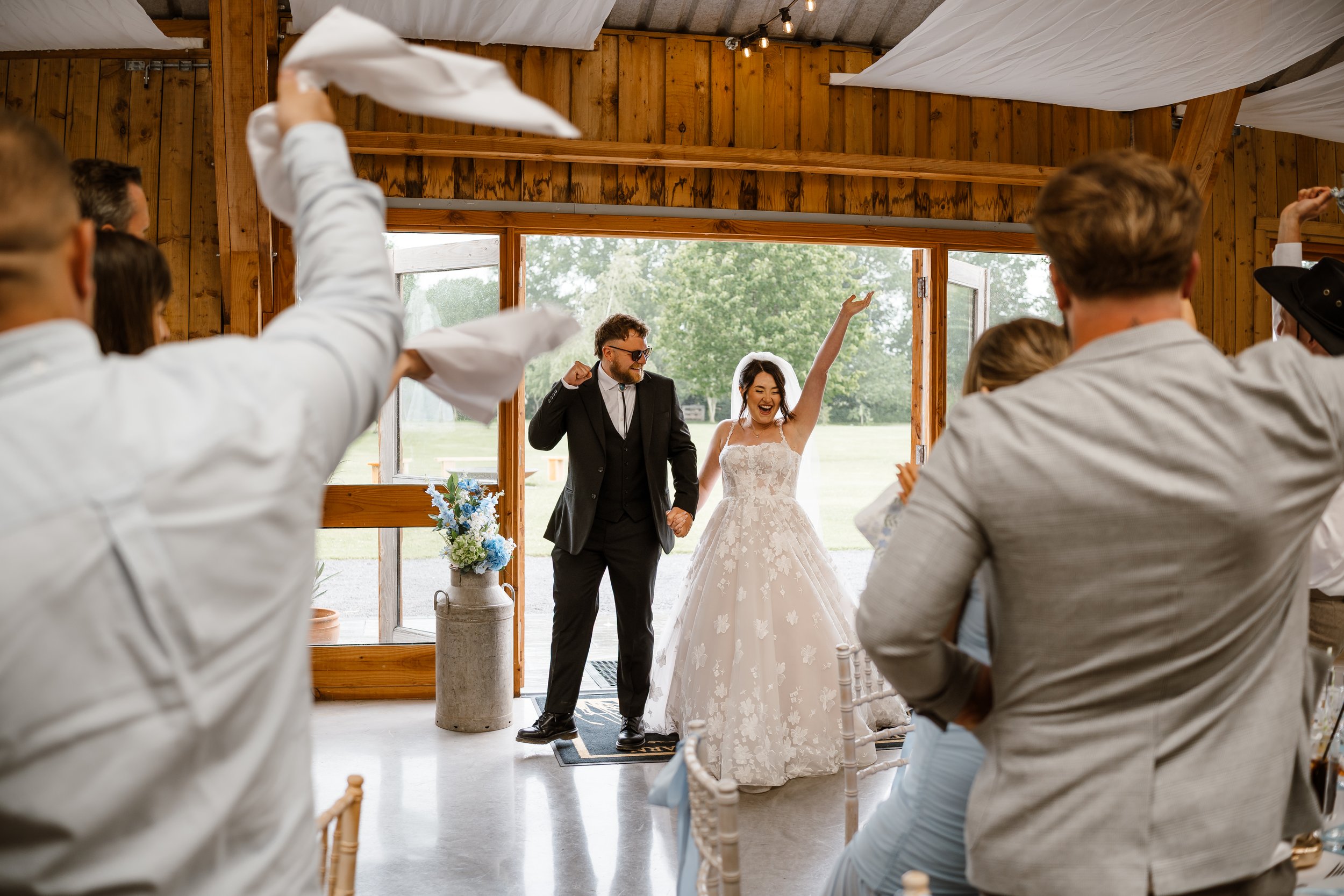 A bride and groom holding hands and smiling at each other during their wedding ceremony inside a rustic wooden venue, with guests seated and celebrating.