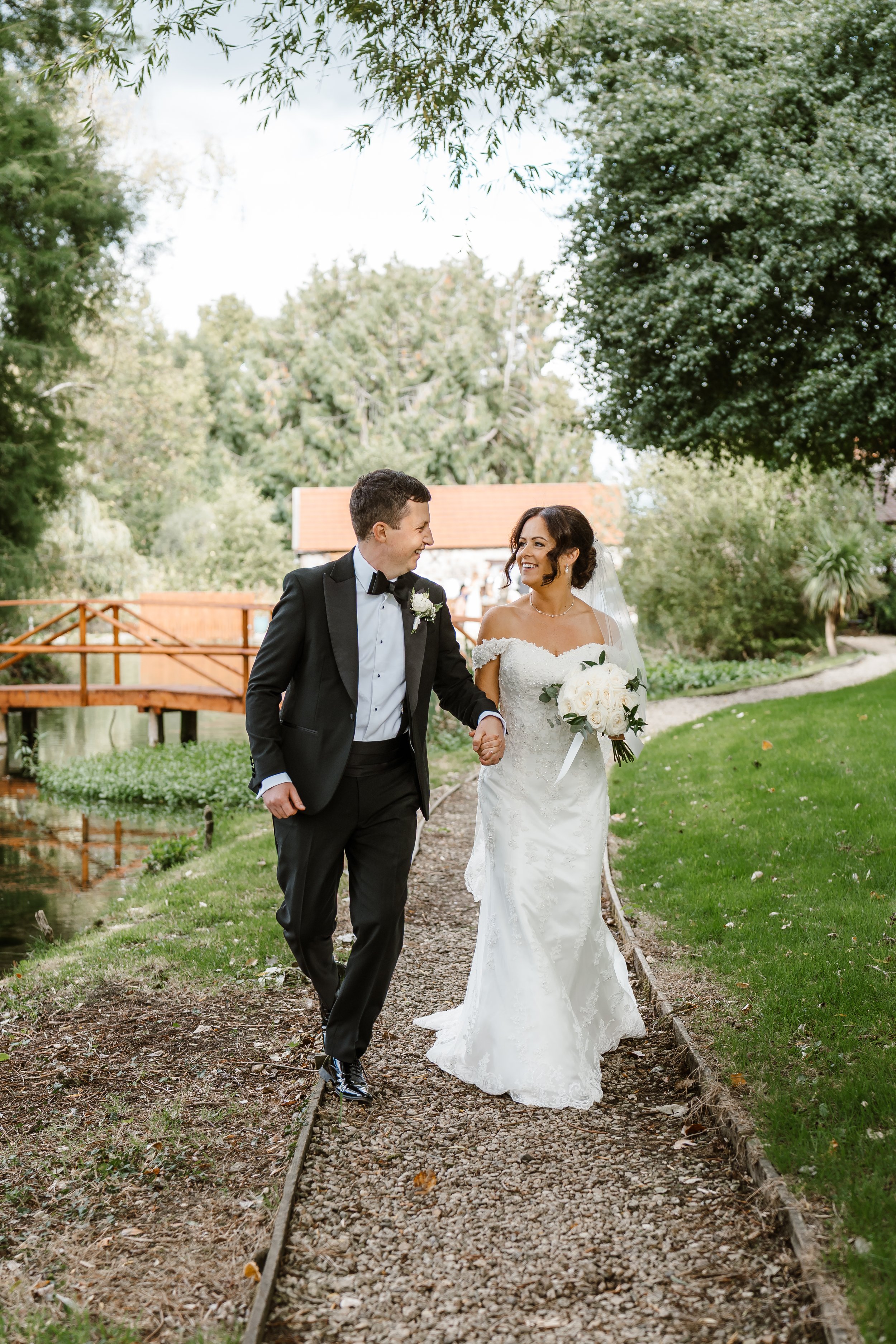A newlywed couple walking along a garden path, holding hands and smiling at each other. The bride is wearing a white off-the-shoulder wedding gown and holding a bouquet of white roses. The groom is dressed in a black tuxedo with a bow tie. The scene 