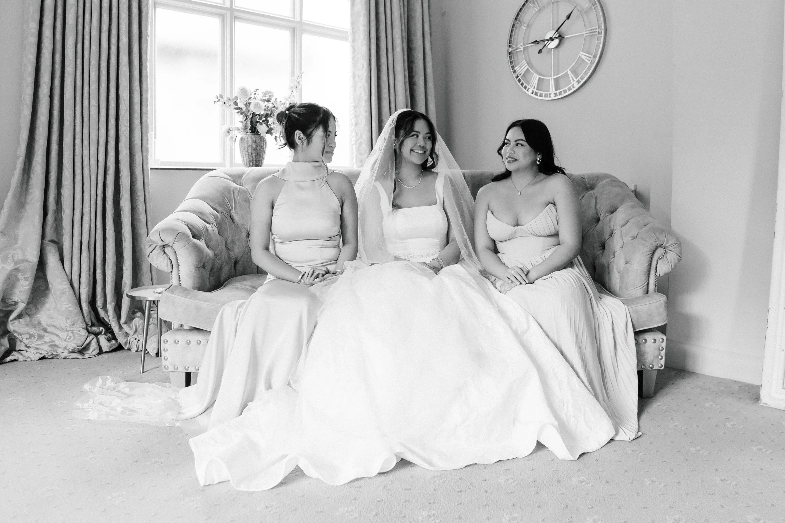 Three women sitting on a sofa in a room with a window and curtains, a floral vase, and a large clock on the wall. The woman in the middle is dressed as a bride in a wedding gown and veil, flanked by two women in evening dresses.