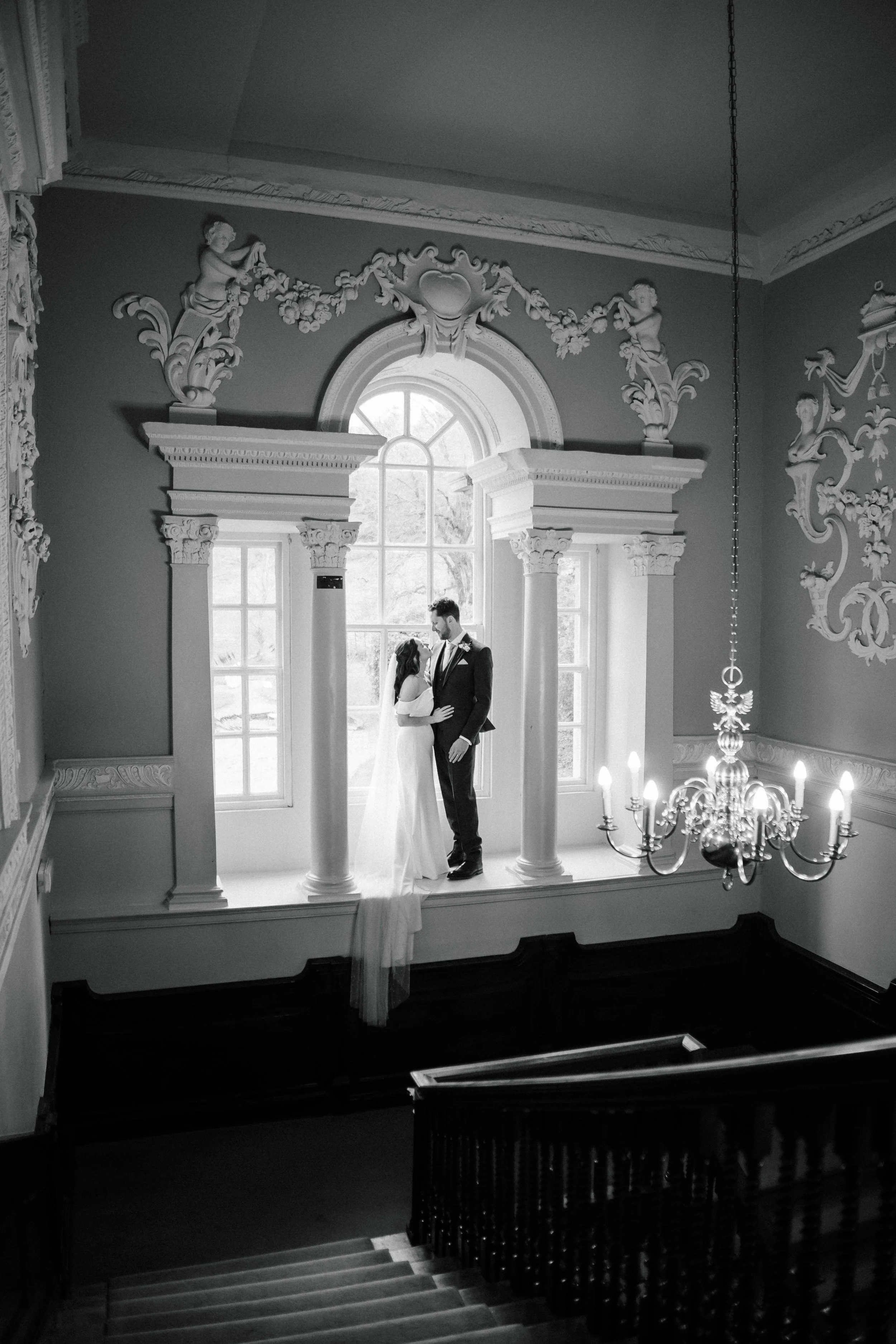 Black and white photo of a bride and groom standing on a window ledge in an ornate room. The bride wears a wedding dress with a long veil, and the groom wears a suit. The room features decorative molding, columns, a chandelier, and a large arched win