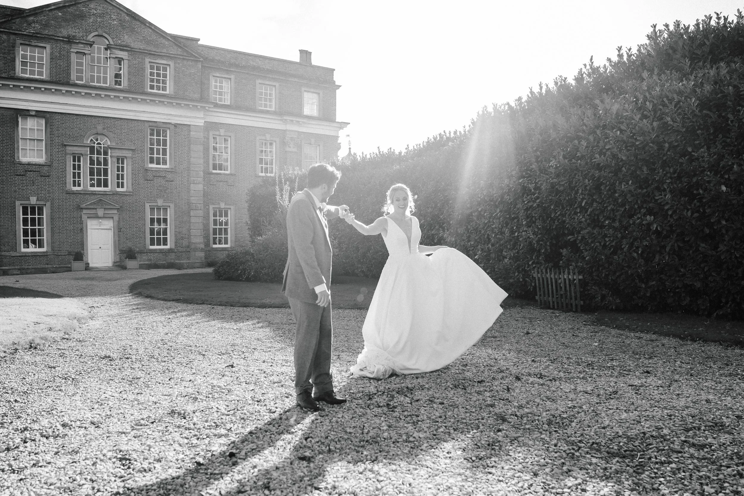 A bride and groom holding hands outside on a gravel path in front of a large brick house. The bride is in a white wedding gown and the groom in a suit. The sunlight is shining brightly, creating long shadows.