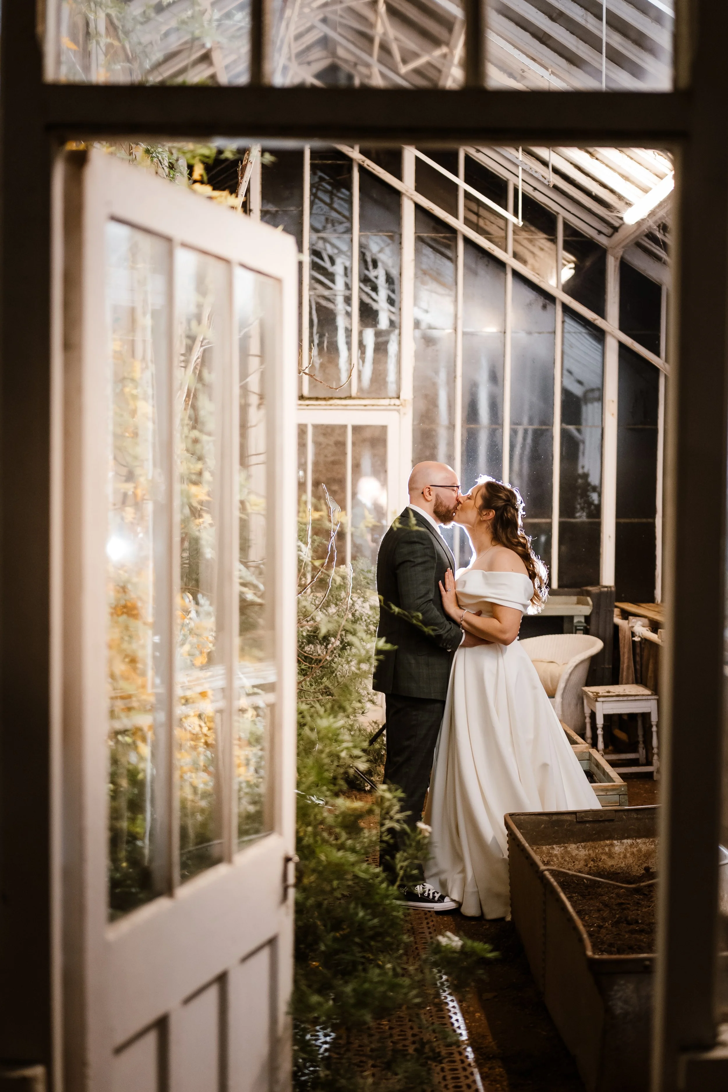 A bride and groom sharing a kiss inside a greenhouse-style structure, visible through a doorway.