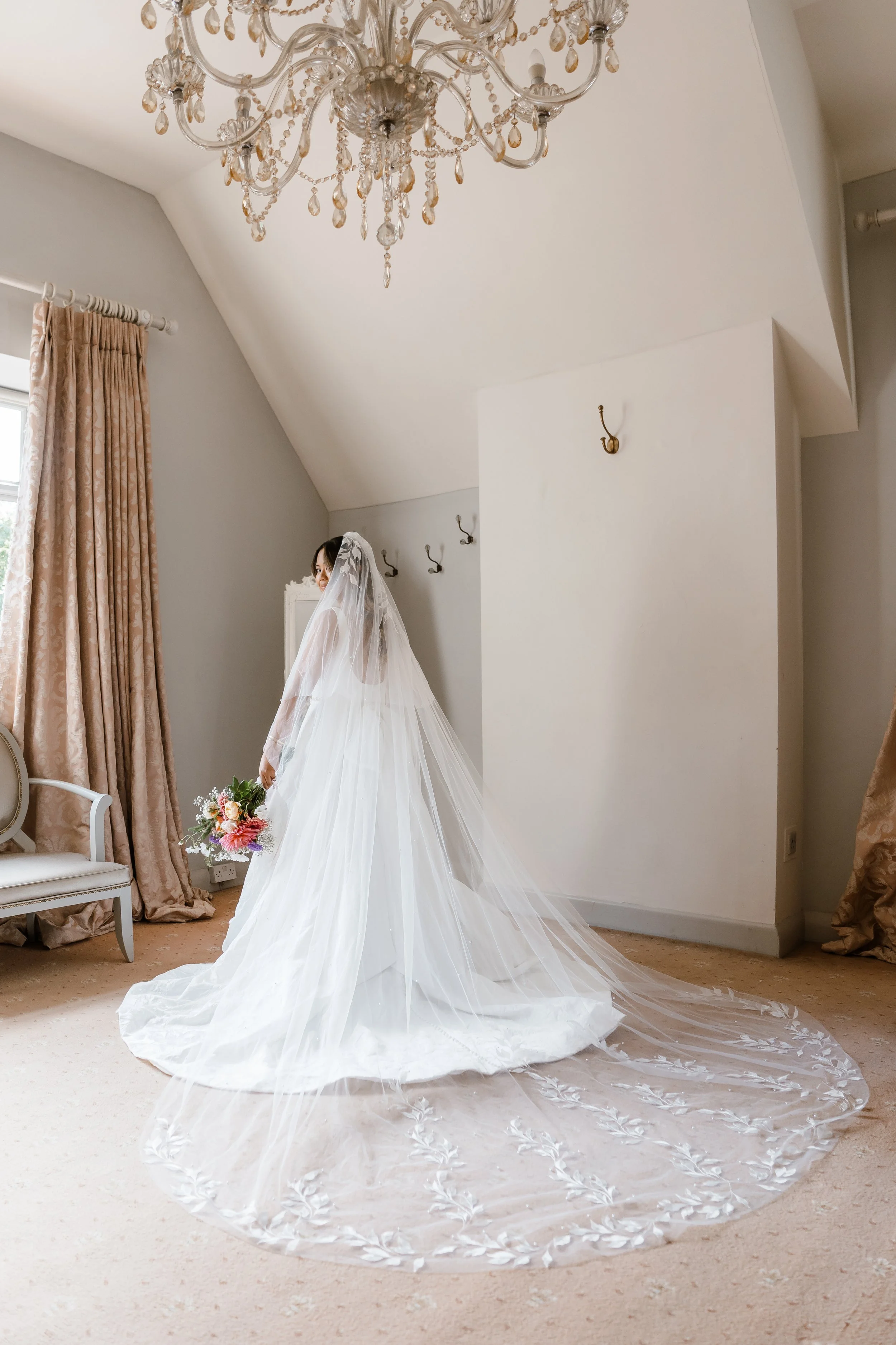 Bride in a wedding dress with a long veil holding a bouquet, standing in a room with beige carpet, pink curtains, and a chandelier.