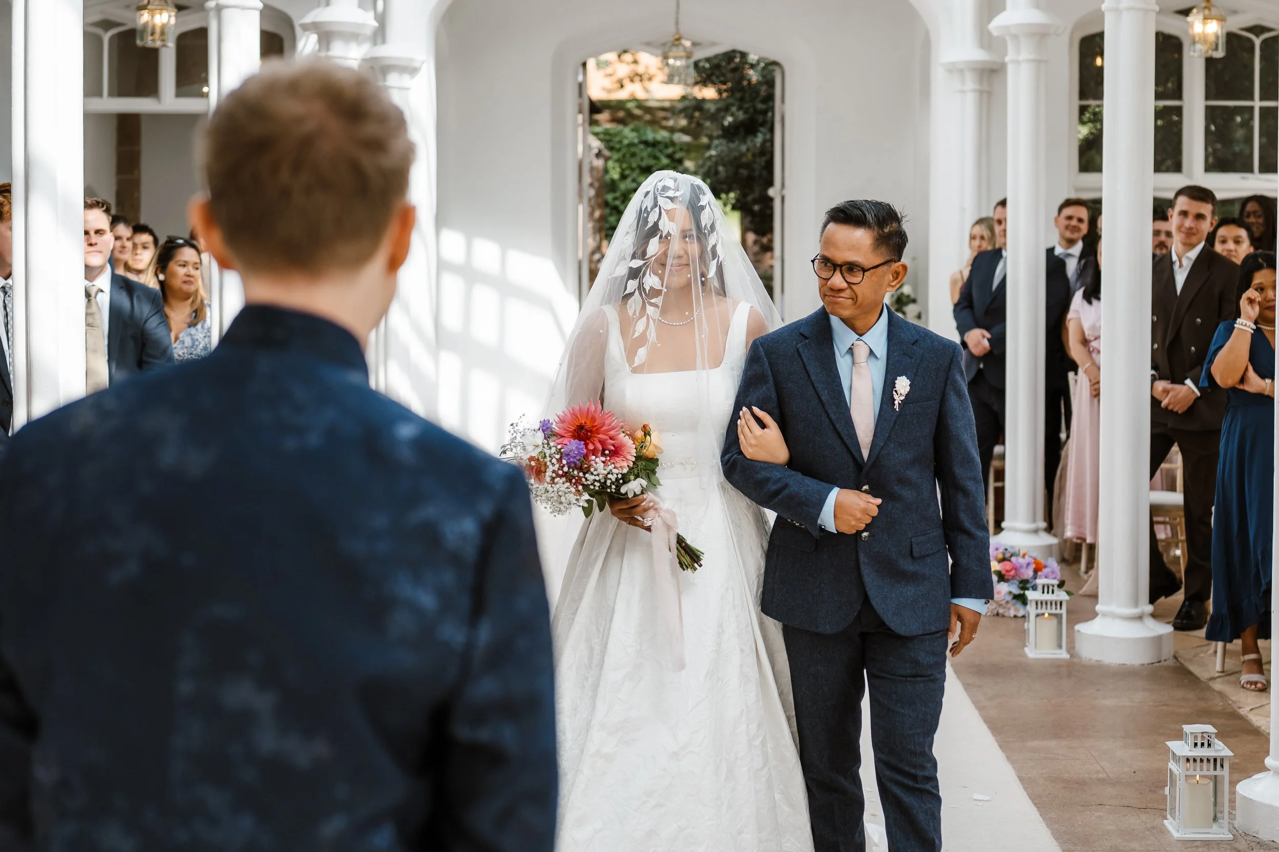 A bride in a white wedding dress with a veil being walked down the aisle by a man in a dark blue suit. The groom, in a dark suit, is facing them, and guests are watching inside a bright, white wedding venue with white columns and lanterns.