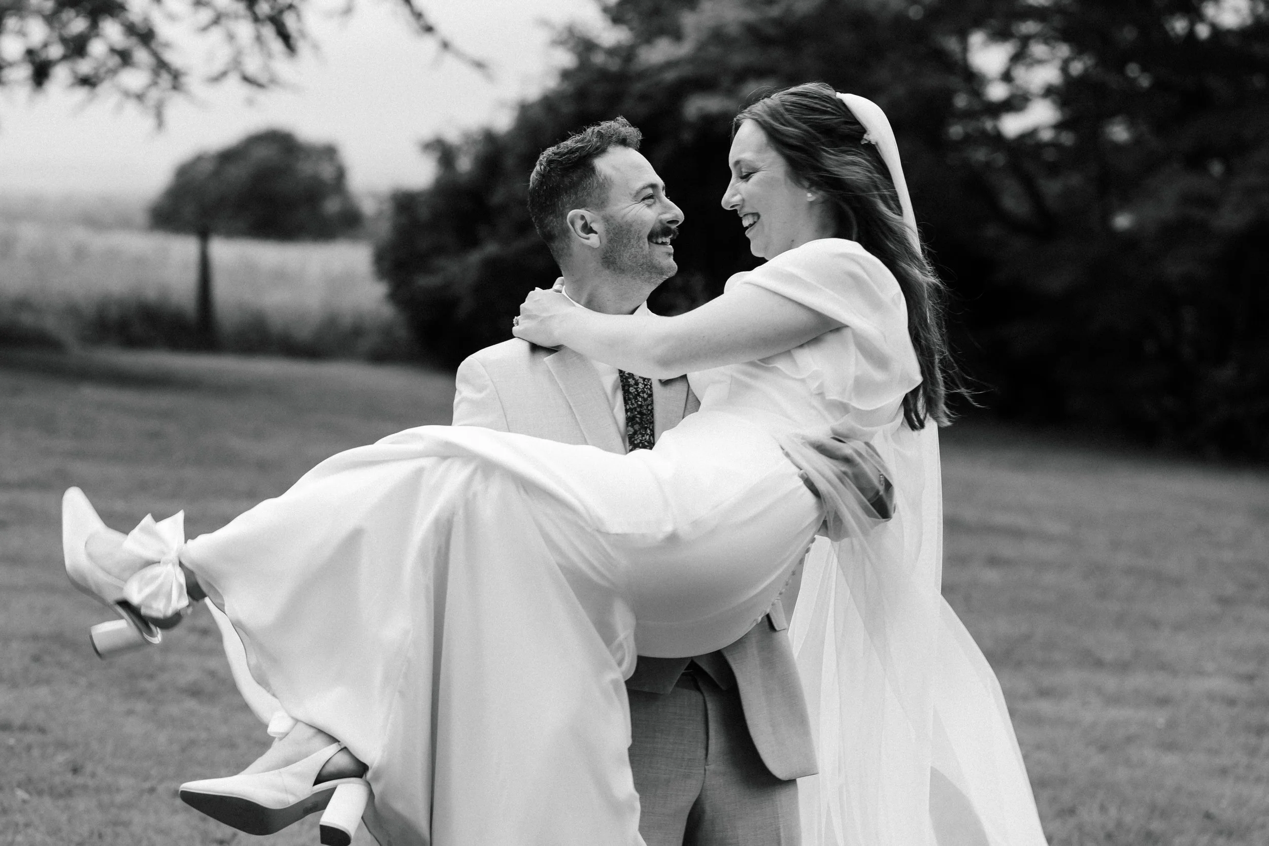 A man in a light-colored suit holding a woman in a white dress, both smiling and happy, outdoors with blurred trees and grass in the background.