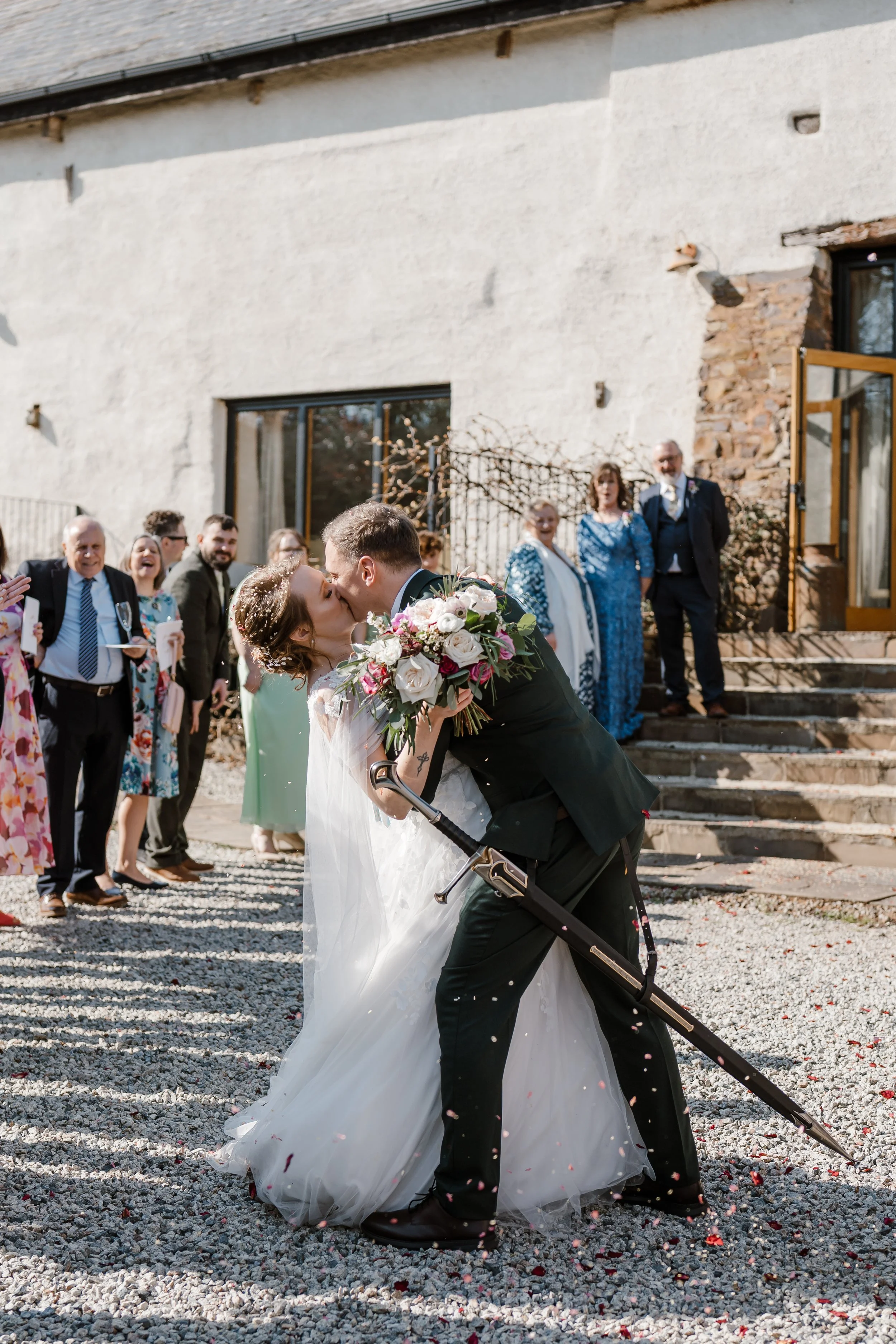 A newlywed couple shares a kiss outside a rustic building as guests celebrate. The bride is in a white wedding gown holding a bouquet, and the groom in a dark suit with a cane. Friends and family stand behind them, smiling and clapping, on a gravel p