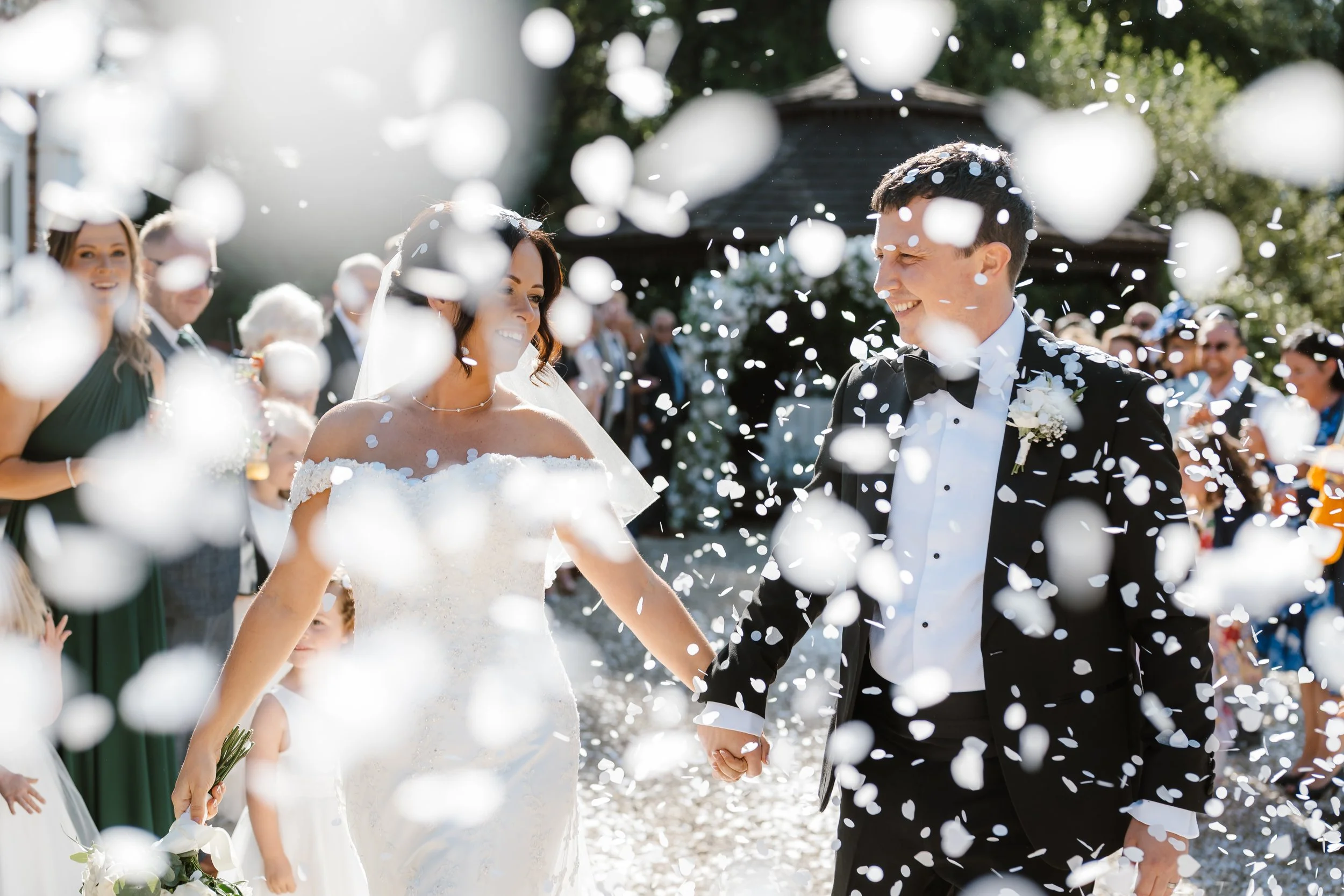 A bride and groom holding hands and smiling during their wedding celebration, surrounded by family and friends, with confetti falling around them outdoors on a sunny day.