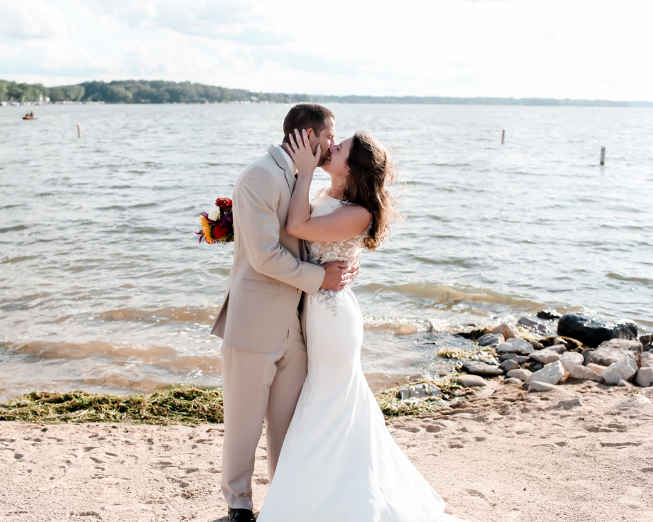 Bride and groom kissing on the sand with the lake in the background at Pewaukee Beach in Pewaukee Wisconsin - Planned by Milwaukee Wedding Planner Natural Elegance LLC and Photos Taken by Memory Lane Photography.png