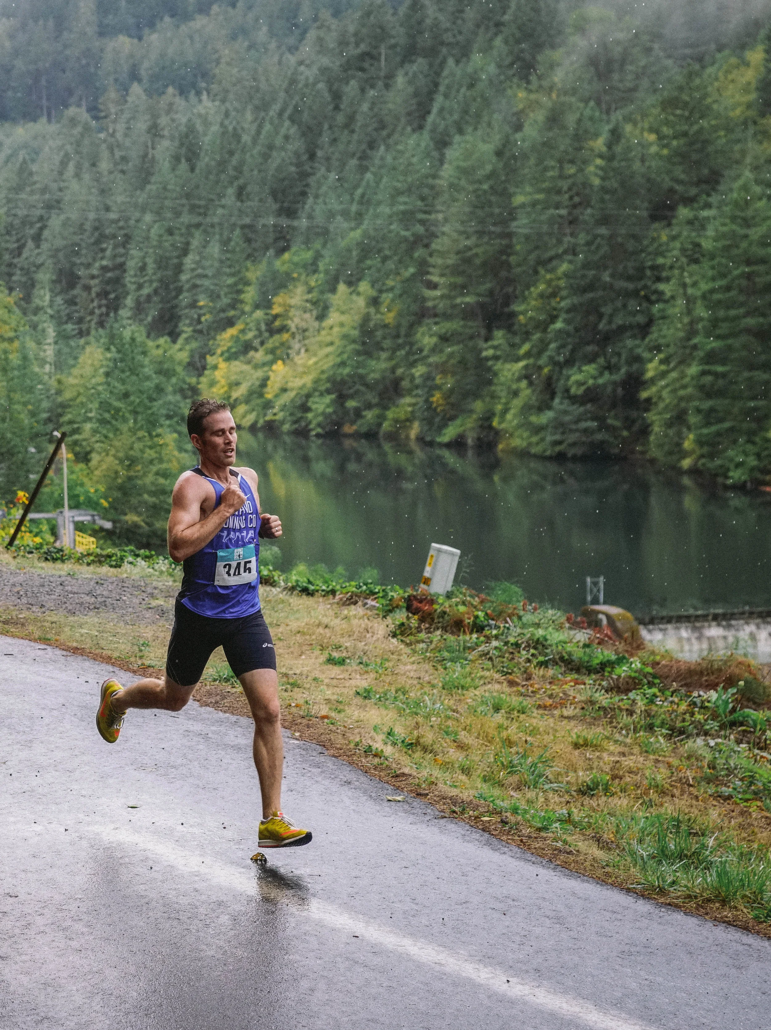 A male runner wearing a blue sports singlet and black shorts running on a paved path beside a lake in Oregon, surrounded by green trees with water reflections.