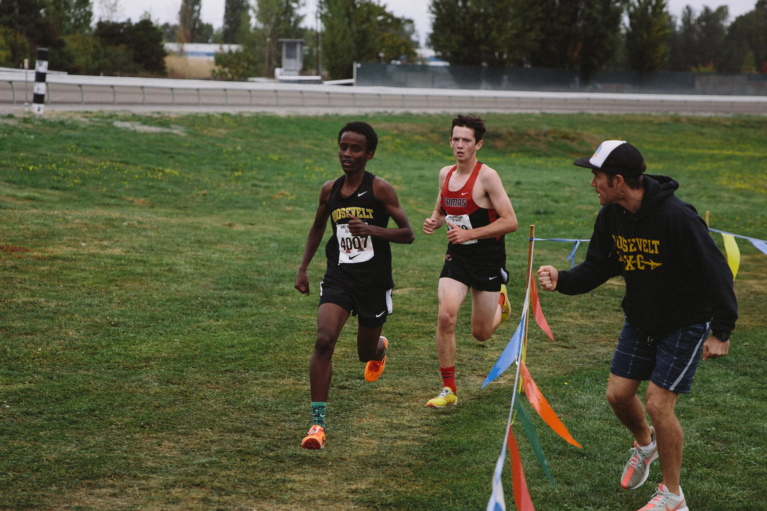Two male runners competing in a race, with one runner wearing a black Roosevelt athletic uniform and the other wearing a red Camas uniform, on a grassy outdoor track. A man is running on the grass beside the runners, guiding and cheering them on.