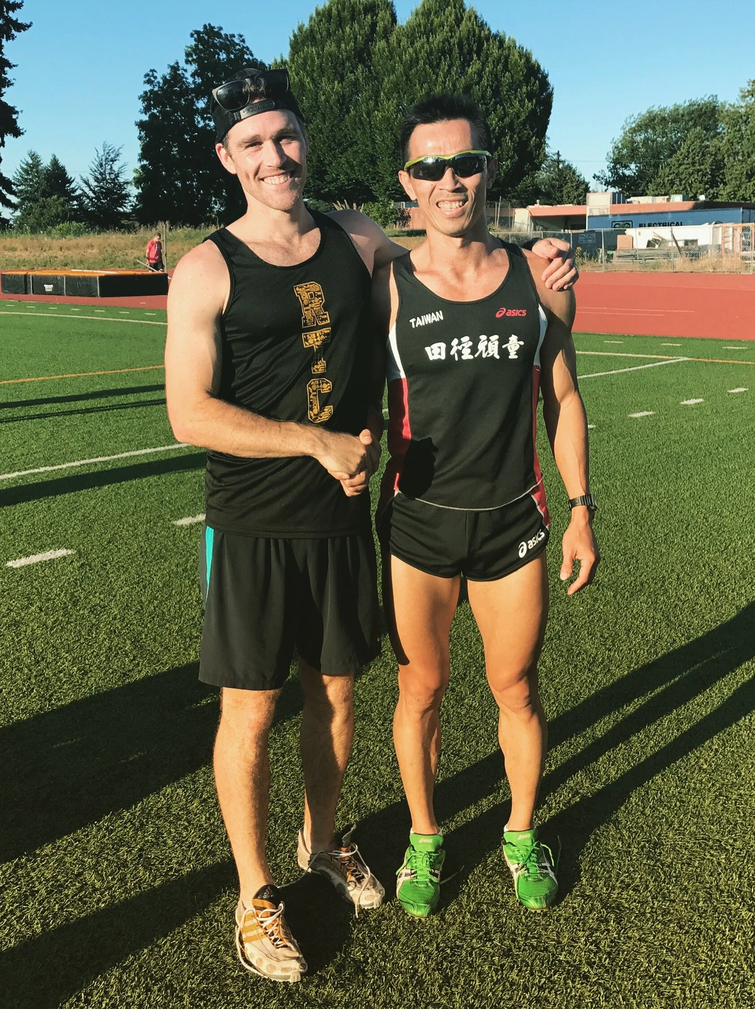 Two men in running athletic attire standing on a track field, smiling and posing for the photo, with trees and a building in the background on a sunny day.