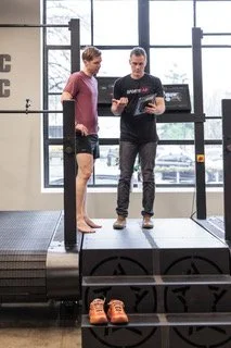 Two men standing on a platform at a gym, looking at a tablet held by one of them. The gym has large windows and exercise equipment in the background.