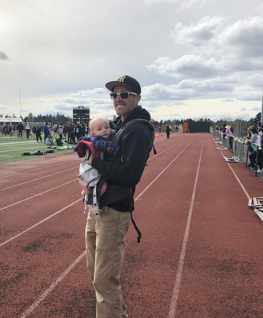 A man holding a baby on a running track at an outdoor sports event with spectators and athletes in the background.
