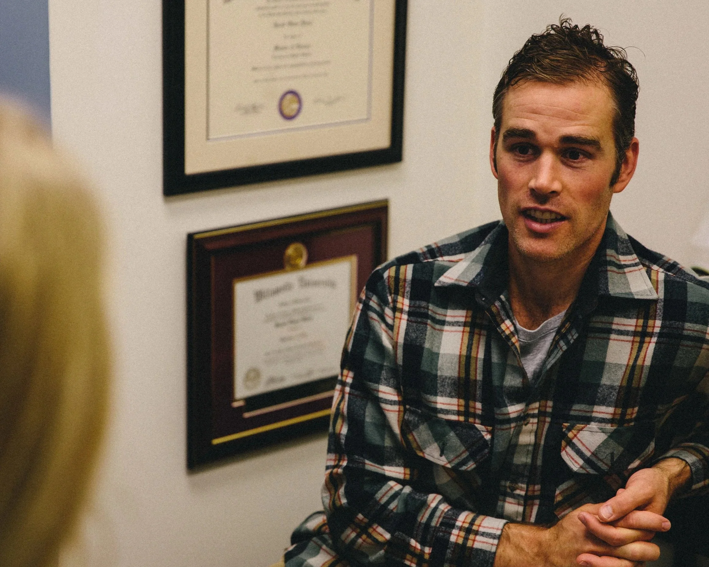 A man with dark, curly hair wearing a plaid shirt is sitting and talking, with framed physical therapy degrees, awards and certificates on the wall behind him.