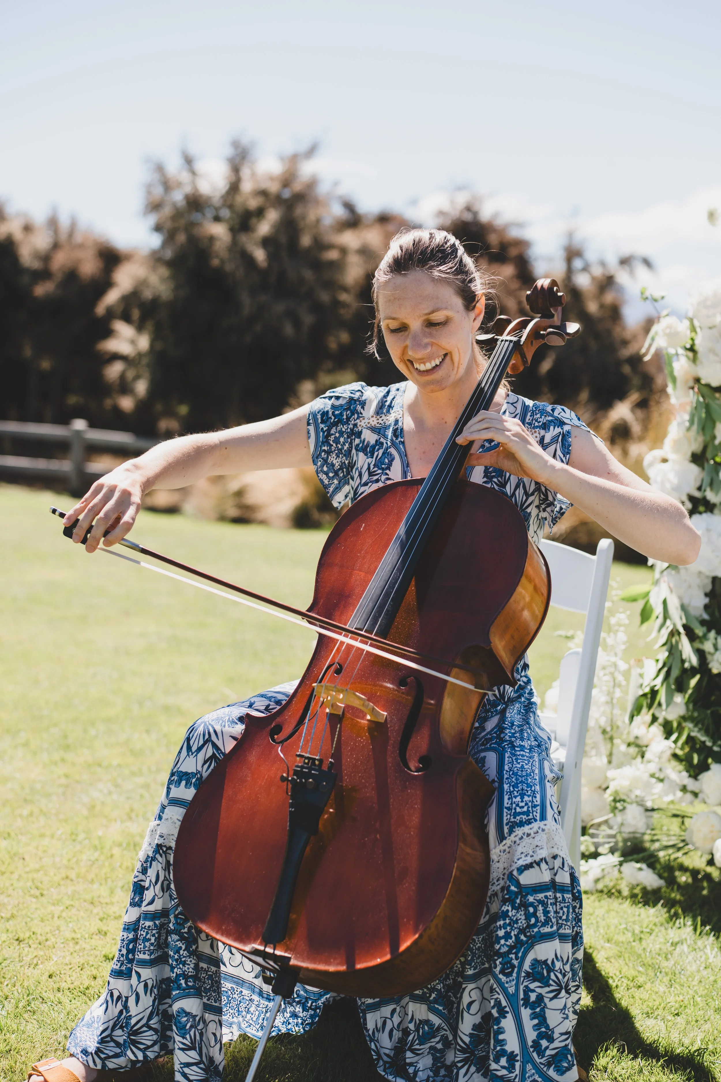 A woman playing a cello outdoors near a lake with mountains in the background during daytime, accompanied by a seated man in formal attire.