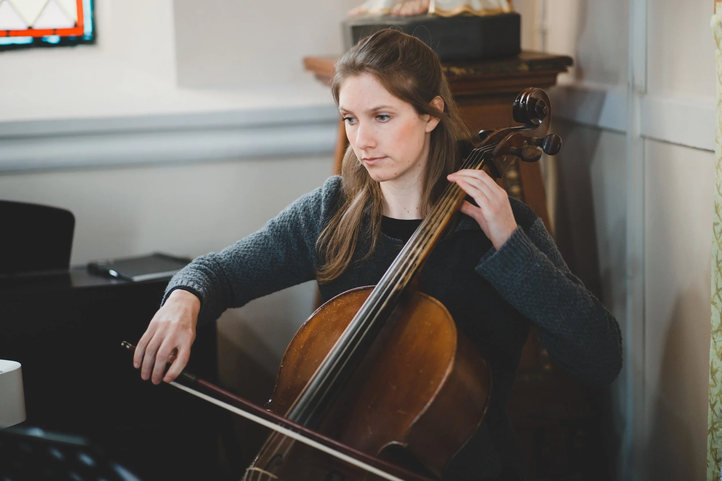 A woman playing a cello outdoors near a lake with mountains in the background during daytime, accompanied by a seated man in formal attire.