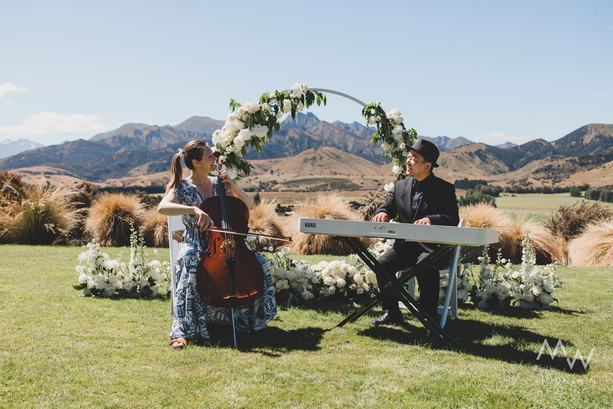 A woman playing the cello and a man playing the keyboard inside a church or chapel with a large arched window, adorned with flowers, decorations, and a small sign.