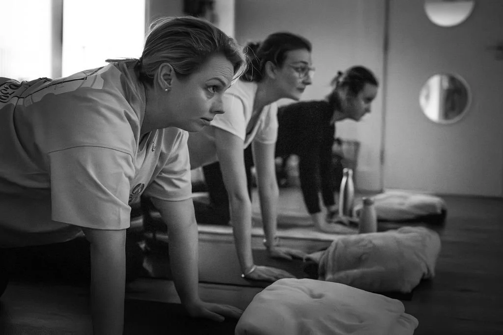 Three women in a yoga class doing a plank pose on mats in a room with circular windows.