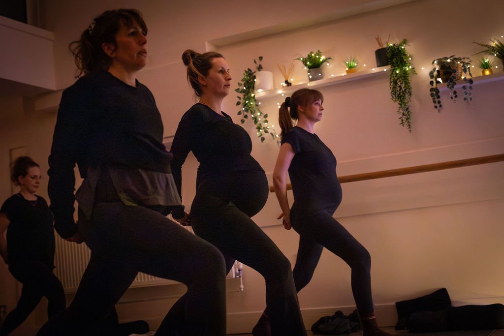 Four pregnant women practicing yoga or dance moves in a dimly lit studio with potted plants and string lights on the wall.