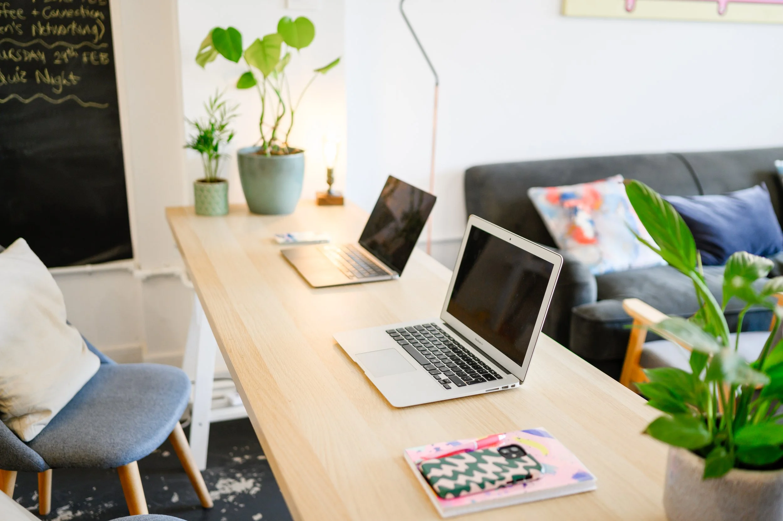 A workspace with two laptops on a wooden table, surrounded by green potted plants, a colorful chair, and a cozy bed in the background.