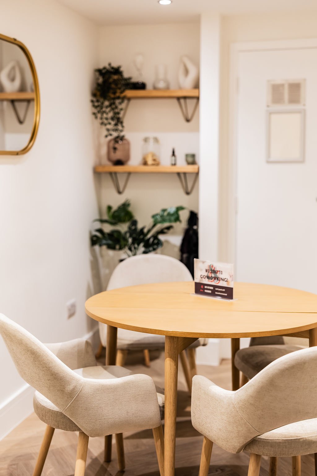 A cozy dining area with a round wooden table surrounded by four upholstered beige chairs in a room with white walls and wooden flooring. In the background, there are two wooden floating shelves with decorative items and potted plants, and a small side table with a potted plant. A mirror with a gold frame hangs on the wall to the left.