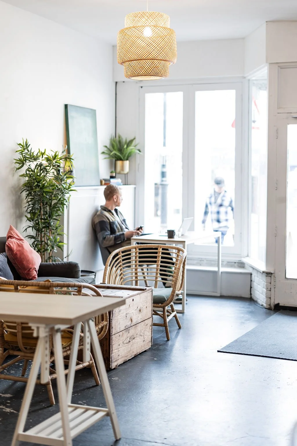 Interior of a cozy cafe with a man sitting by a window using a laptop, surrounded by plants, wooden furniture, and a woven ceiling light.