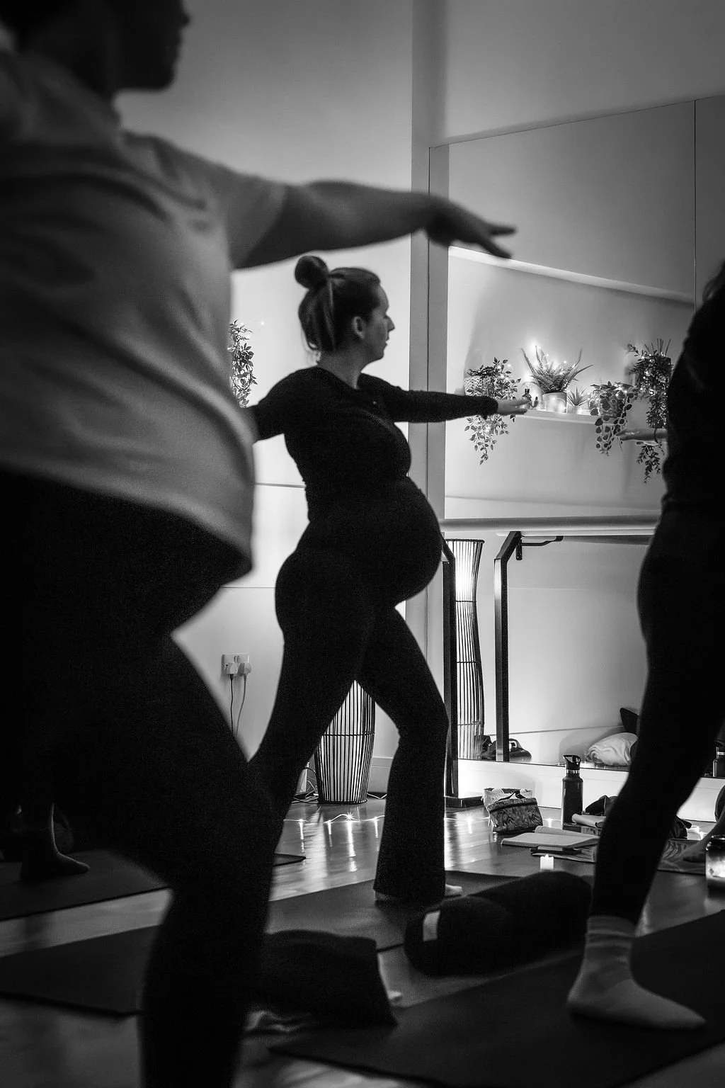 People participating in a prenatal yoga class, with a pregnant woman leading the session, in a room decorated with plants and people practicing yoga poses.
