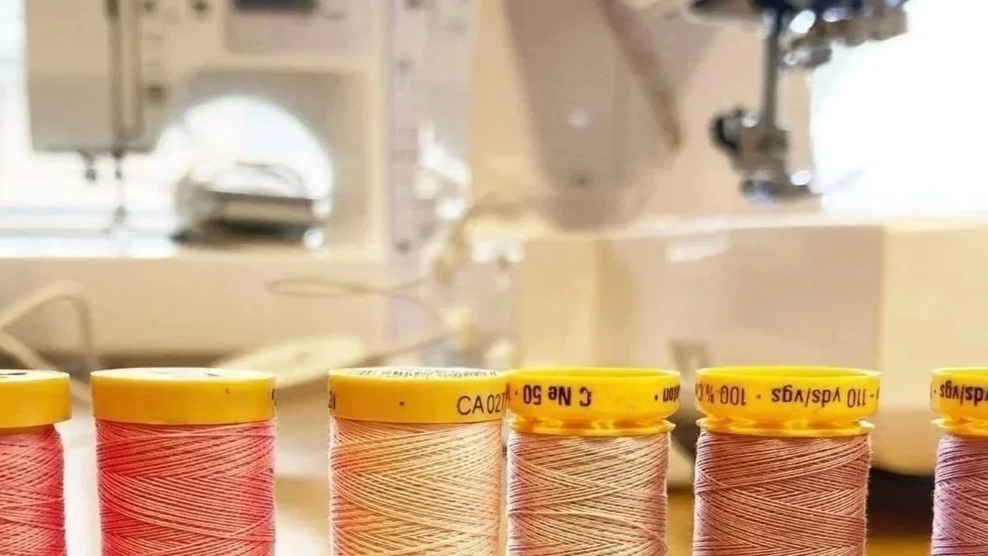 Six spools of pink thread with yellow caps on a sewing table, with a sewing machine and a bowl in the background.