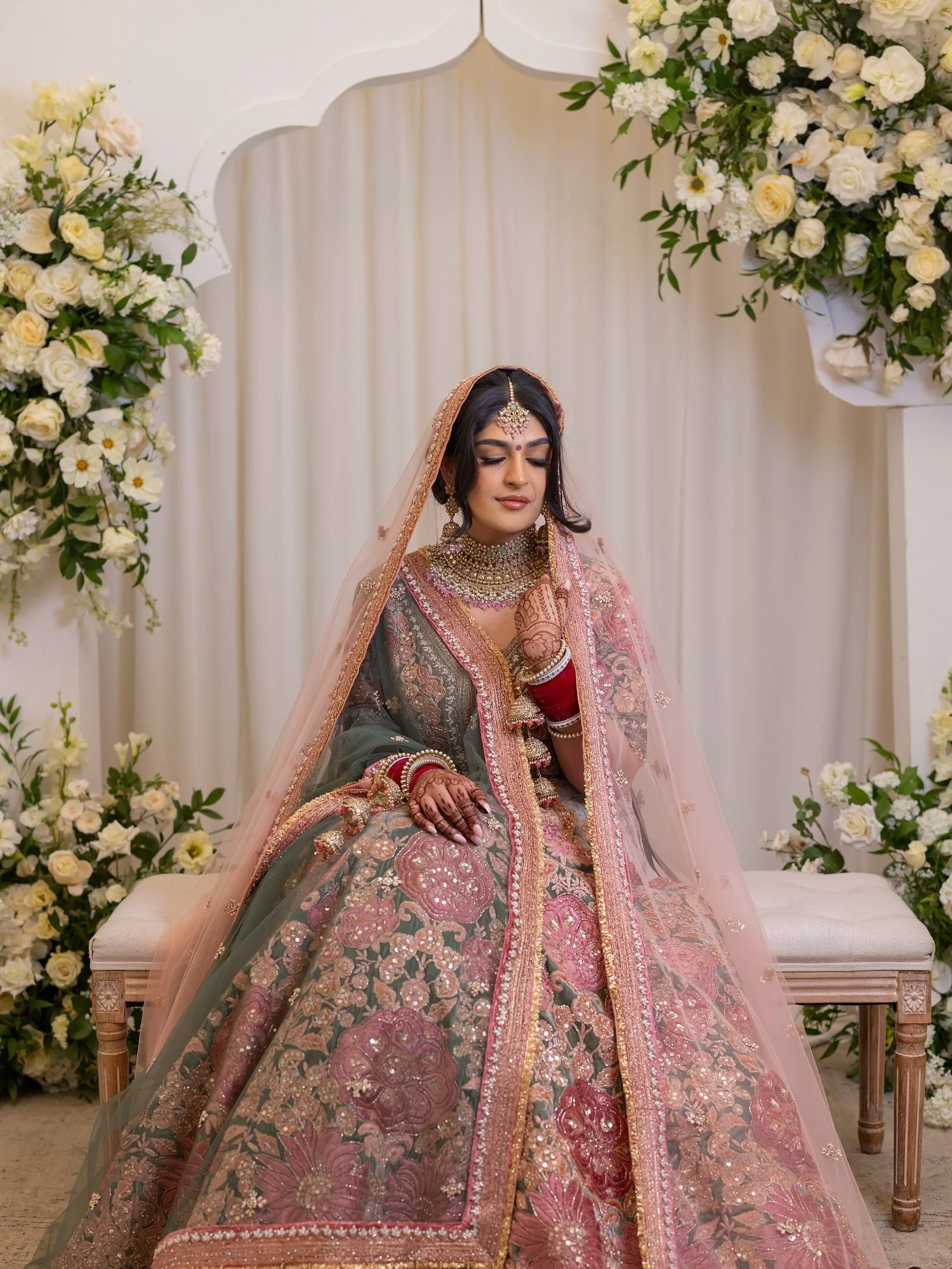 3/3 - Sonya sits in serene splendor, wrapped in the elegance of her intricately embroidered bridal lehenga. The soft blush dupatta, vibrant florals, and detailed mehndi capture the grace, tradition, and joy of a bride glowing from within. This is the
