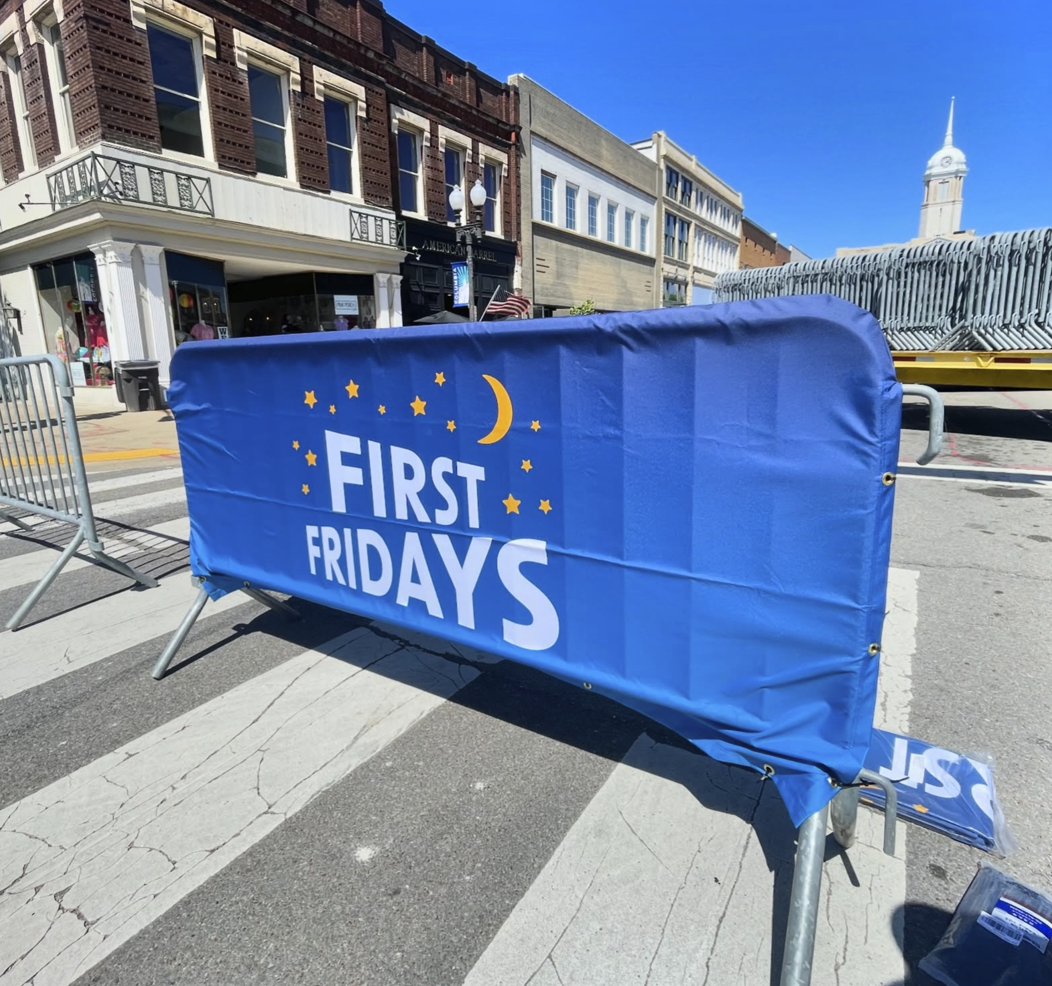 Street barricade with a blue cover displaying the text "First Fridays" and a moon and star design, set up on a city street during daytime.