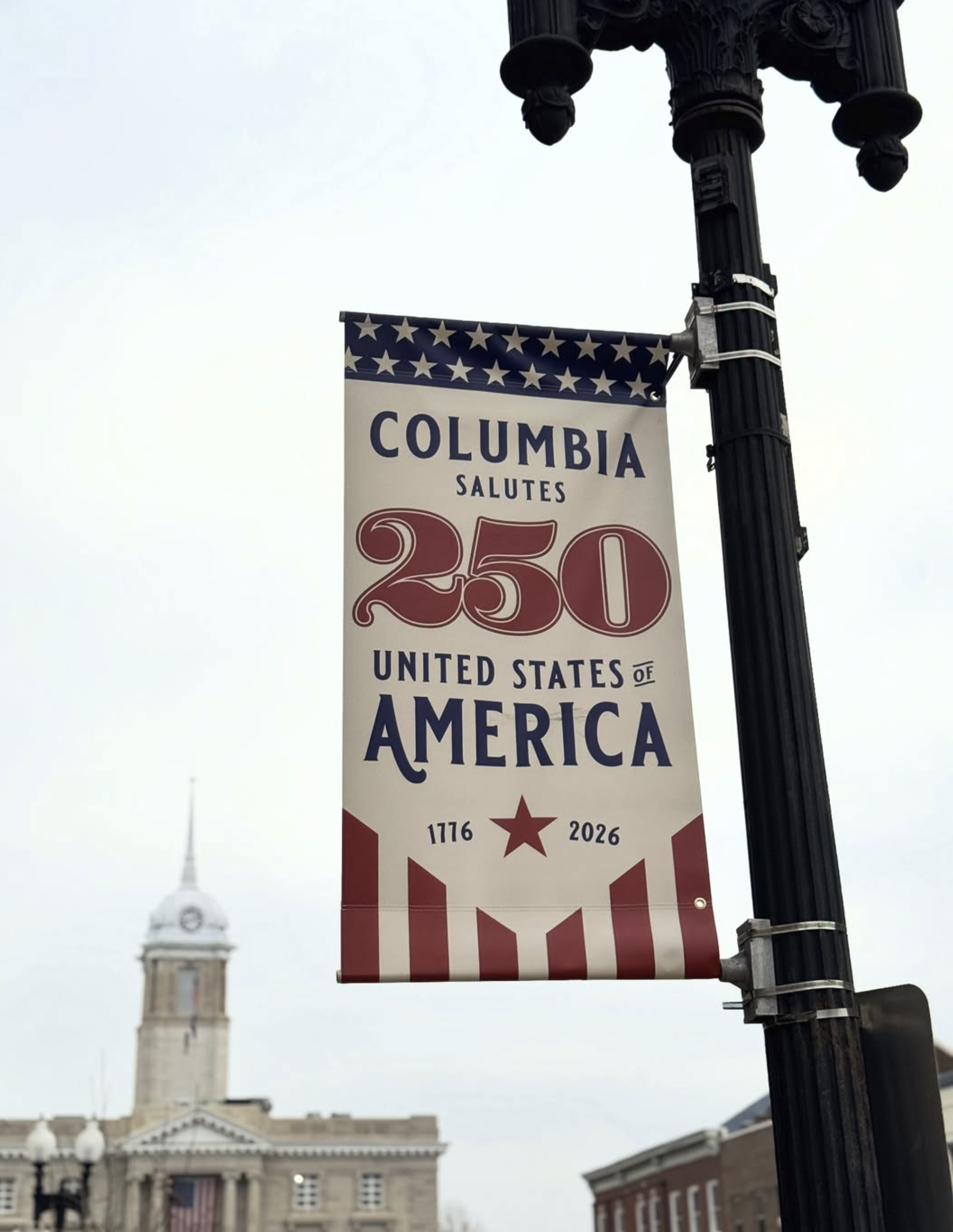A patriotic banner hanging from a streetlamp post with the words 'Columbia Salutes 250' and the dates 1776 and 2026. The banner features stars and stripes in red, white, and blue. In the background, a historic building with a clock tower is visible, with gray overcast skies.