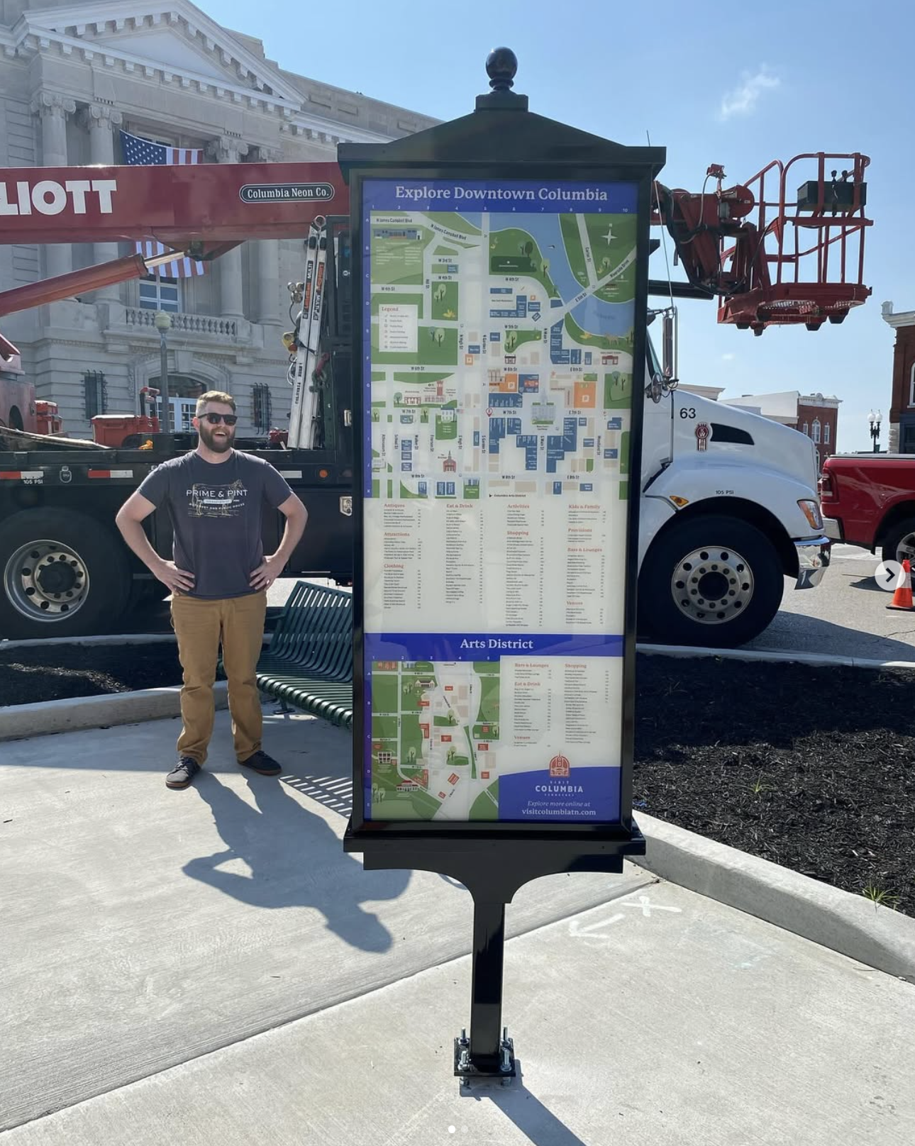 A man stands next to a large city map sign in downtown Columbia, with a construction truck in the background and a historic building with American flags.